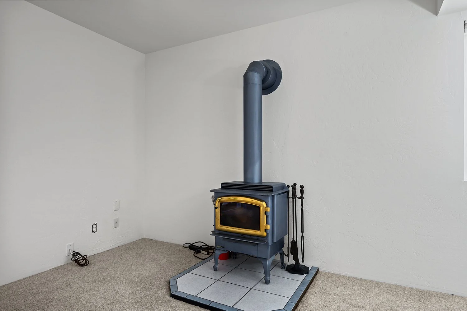 A small blue wood stove with a gold trim and glass door, installed on a tiled hearth in a white-walled room. Black fireplace tools stand next to it, and a black chimney pipe extends upward from the stove to the ceiling.