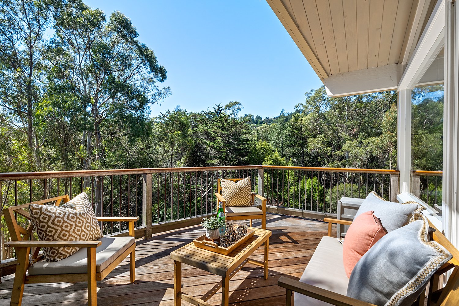 Wooden balcony with outdoor seating including a sofa and chairs with patterned pillows, a coffee table with a tray, bottle, and plant, overlooking lush green trees under a clear blue sky.