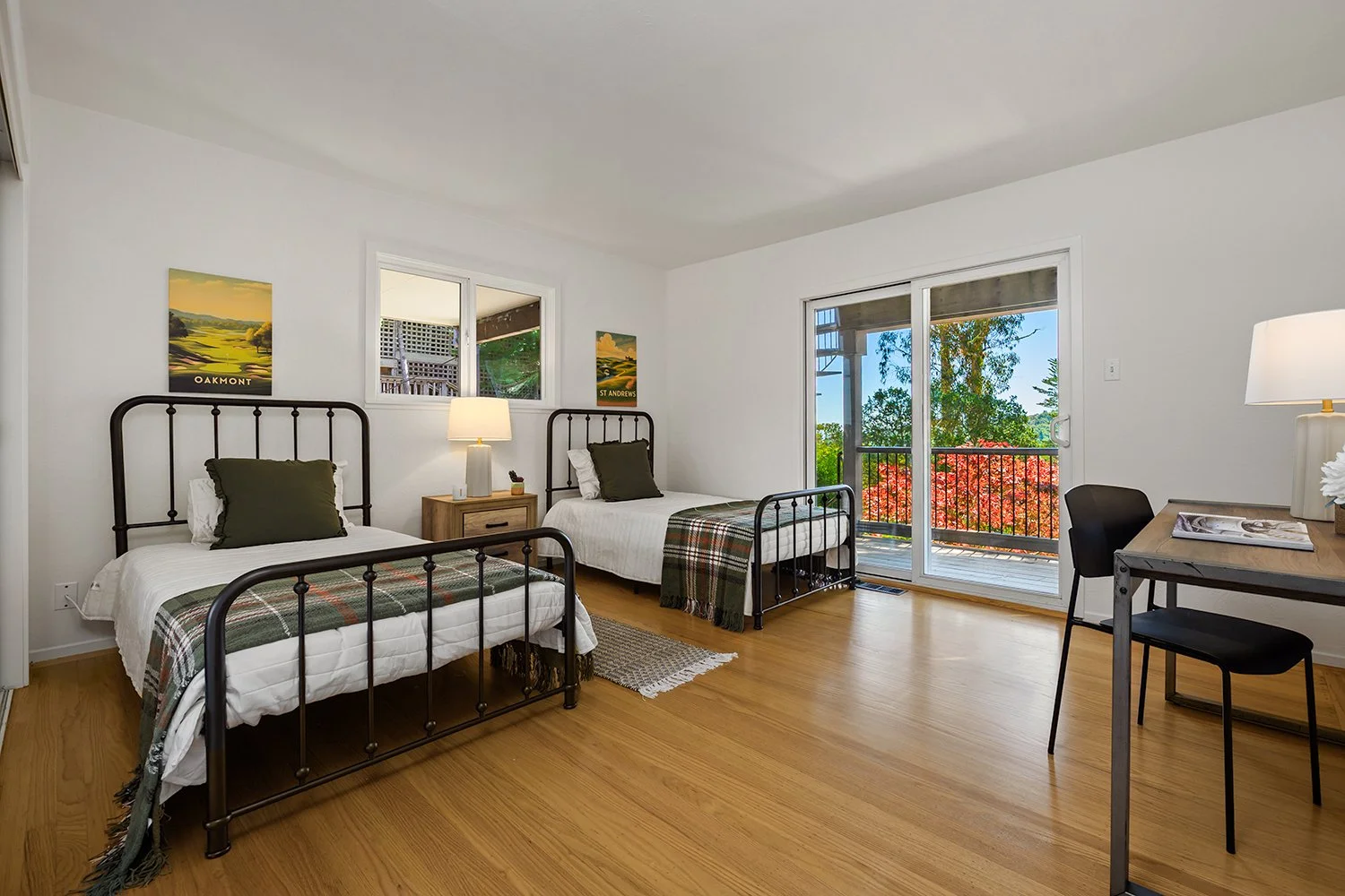 A bright bedroom with two queen beds with black metal frames, white bedding, and dark green pillows. A wooden nightstand with a lamp and small decor is between the beds. A sliding glass door opens to a balcony with greenery and a colorful tree outsid