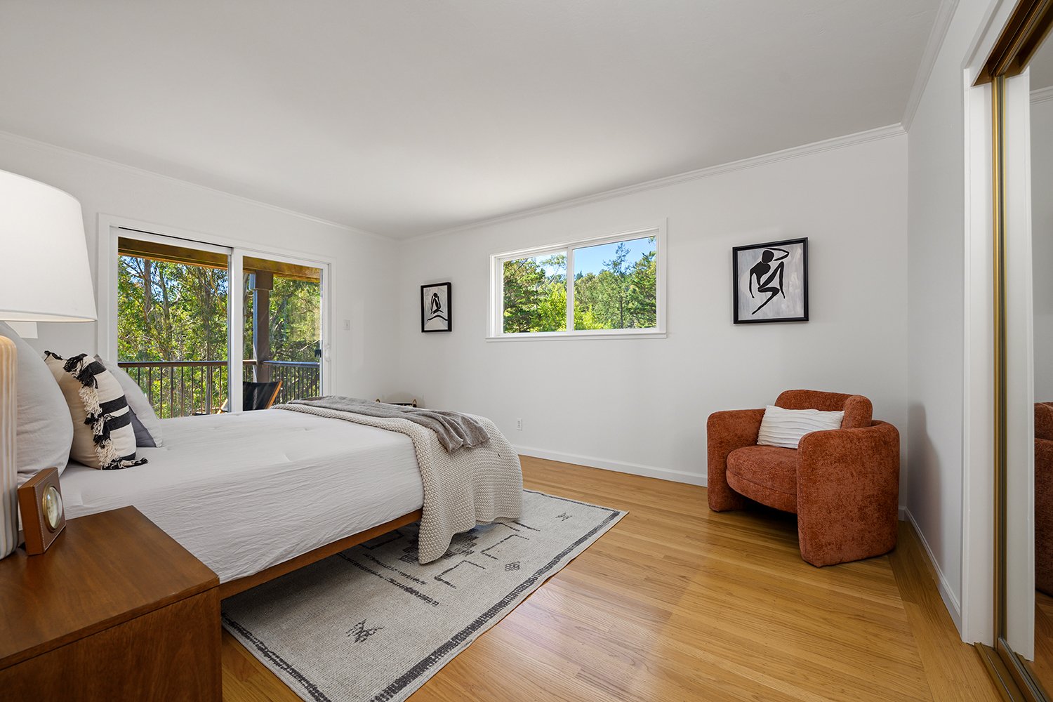 A bedroom with a white bed, wooden nightstand, patterned rug, window and sliding glass door leading to a balcony, orange armchair with a pillow, and framed art on white walls.