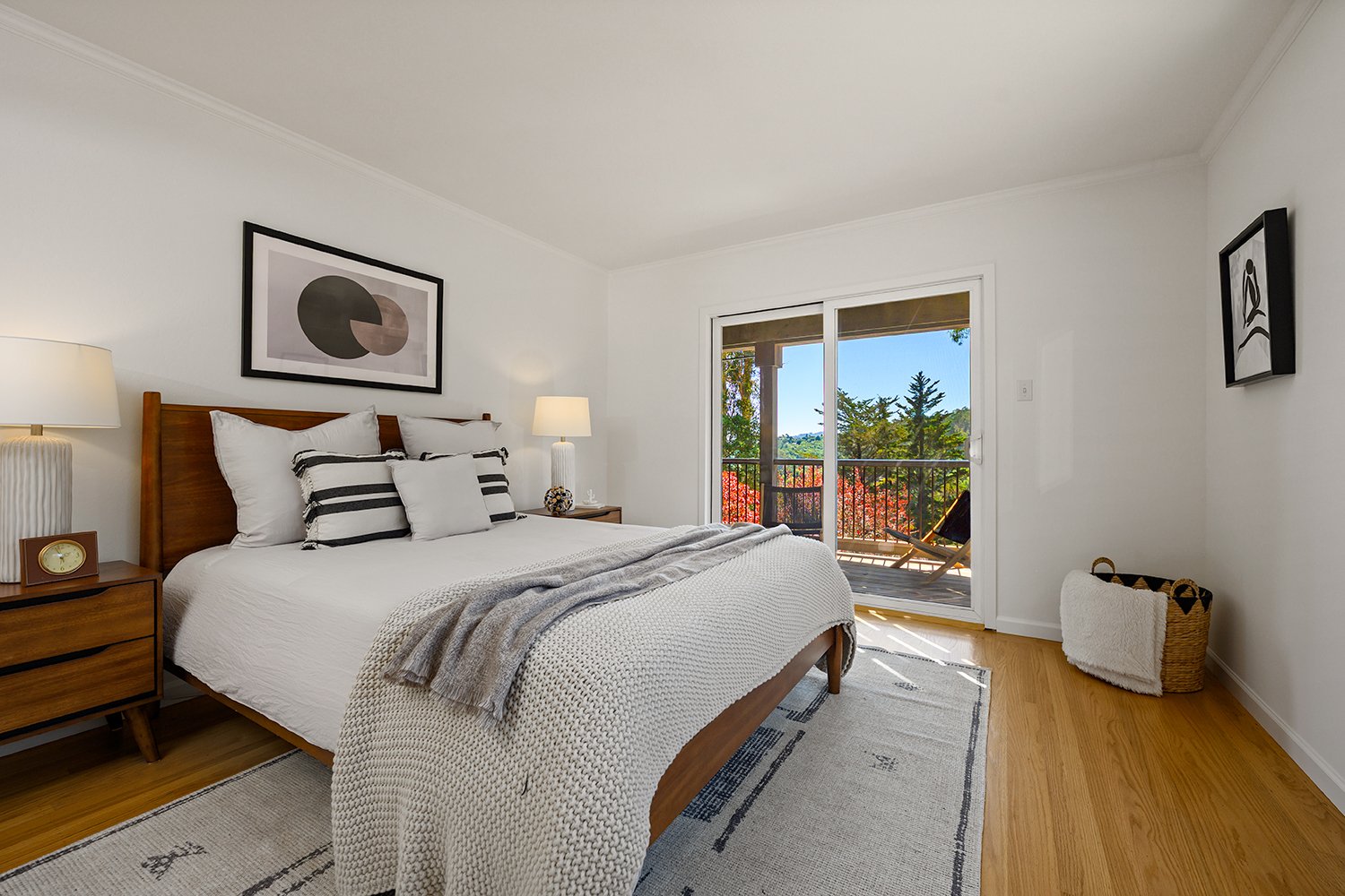 Bedroom with a wooden bed, white bedding, striped pillows, two lamps, outside balcony with chairs, wood flooring, and decor including two framed pictures on white walls.