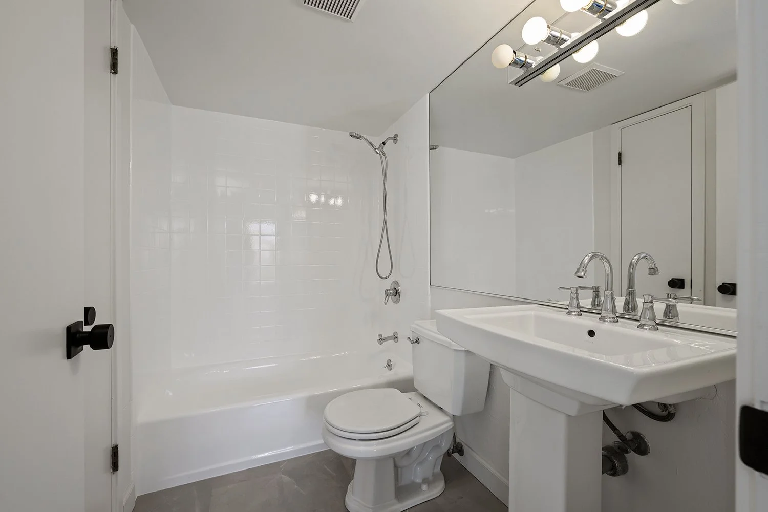 A small modern bathroom with a white bathtub, toilet, and rectangular sink with a large mirror above. The bathroom has a gray floor and white walls, the mirror reflects a light fixture with multiple bulbs.