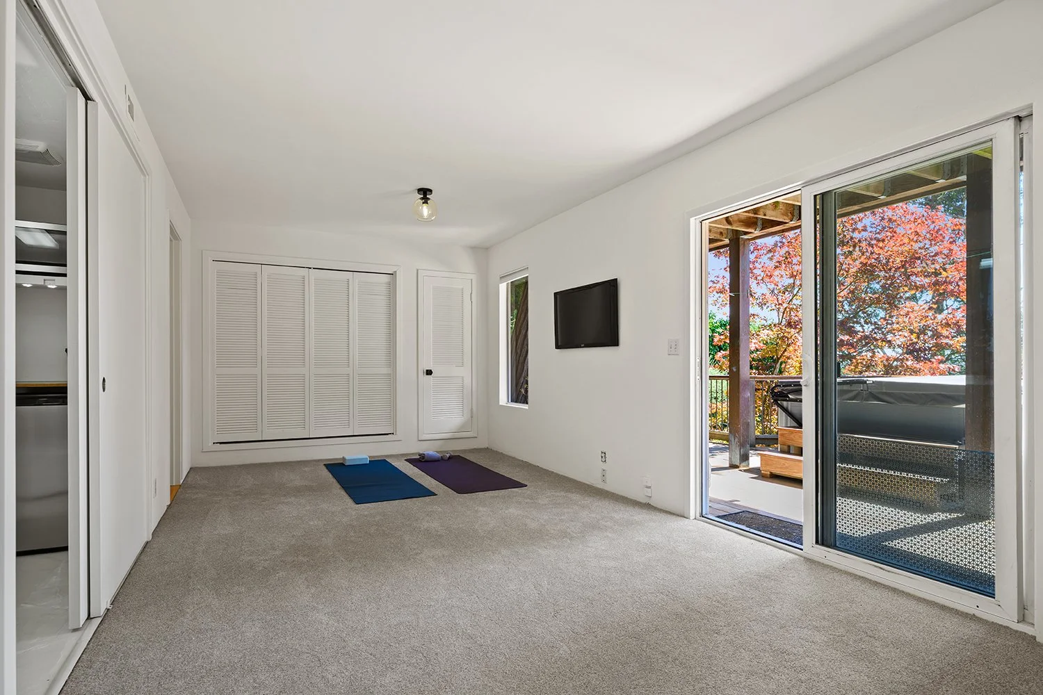 Empty living room with white walls, a flat-screen TV, sliding glass door to a balcony, two yoga mats on carpeted floor, and a closet with louvered doors.