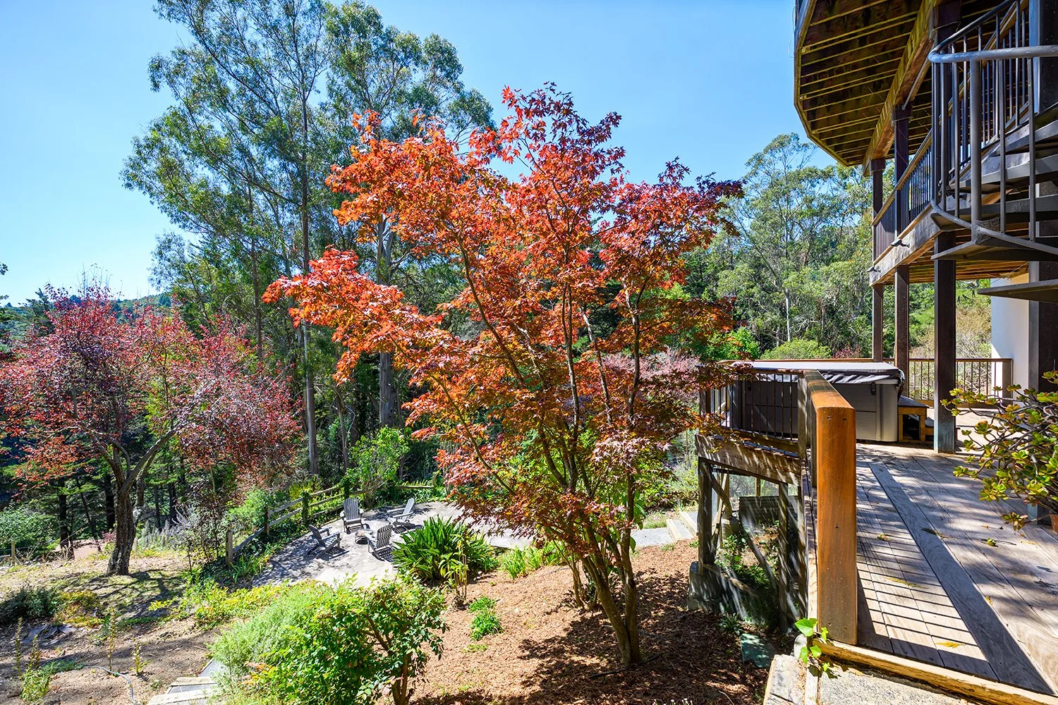 A backyard with colorful trees, wooden deck, hot tub, and outdoor seating area, surrounded by lush greenery.
