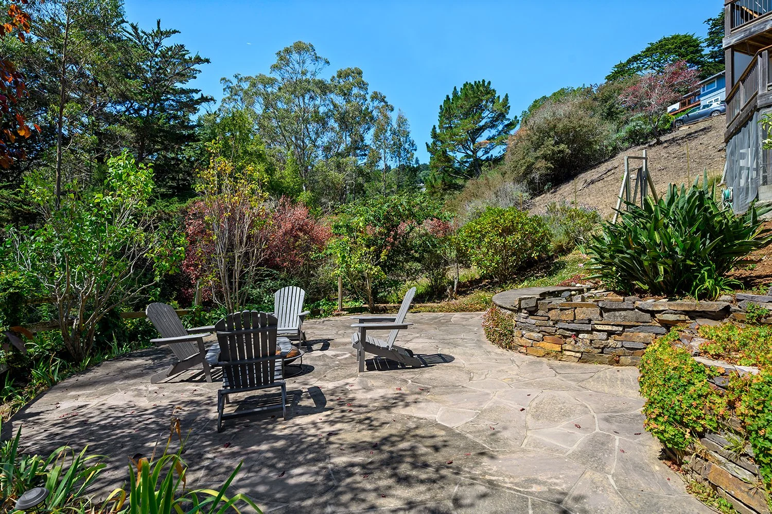 A backyard patio with stone flooring, surrounded by lush green trees and bushes, with four Adirondack chairs arranged in a circle, and a built-up stone garden bed on the right side.