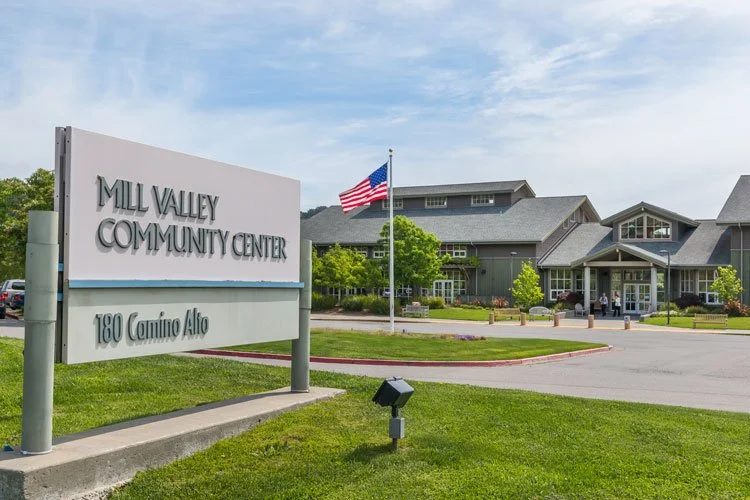 The Mill Valley Community Center with a large sign in the foreground and a building with a gray roof and multiple windows in the background, along with an American flag and green landscaping.