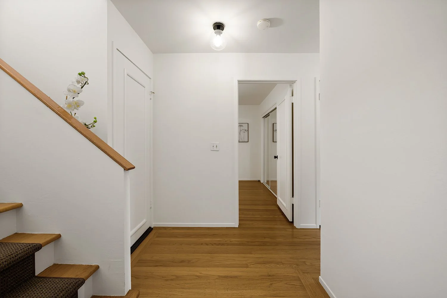 Interior view of a hallway with white walls, hardwood floors, a staircase on the left with a wooden handrail and a flower decoration, and a doorway with mirrored closet doors.