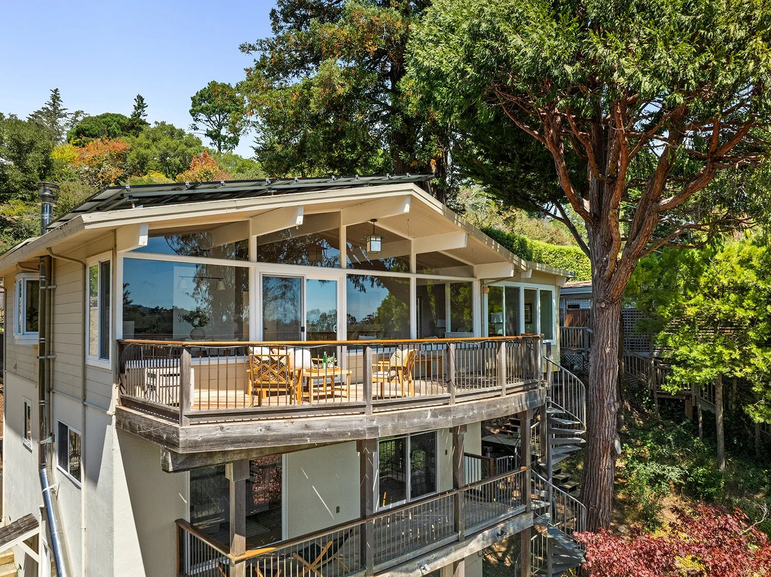Multi-story house with large glass windows, surrounded by tall trees and greenery, featuring a spacious balcony with outdoor furniture.