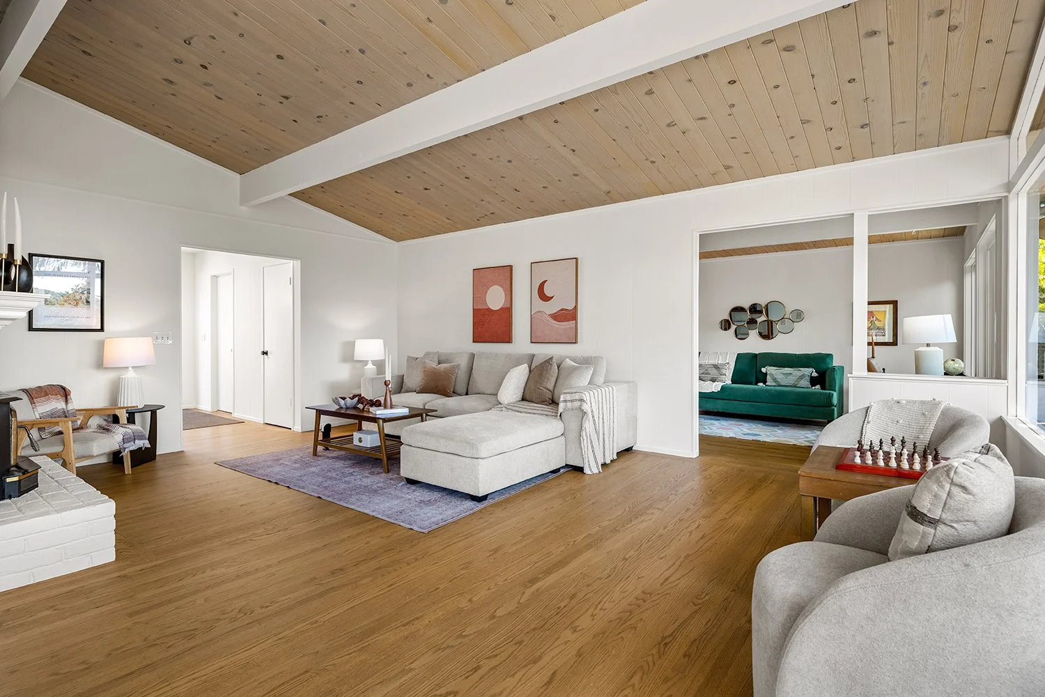 Living room with a high wooden ceiling, white walls, and hardwood floors. Contains a light-colored sectional sofa, a gray armchair with a chessboard, and a small wooden side table. Two abstract art pieces hang above the sofa. A cozy space with large 