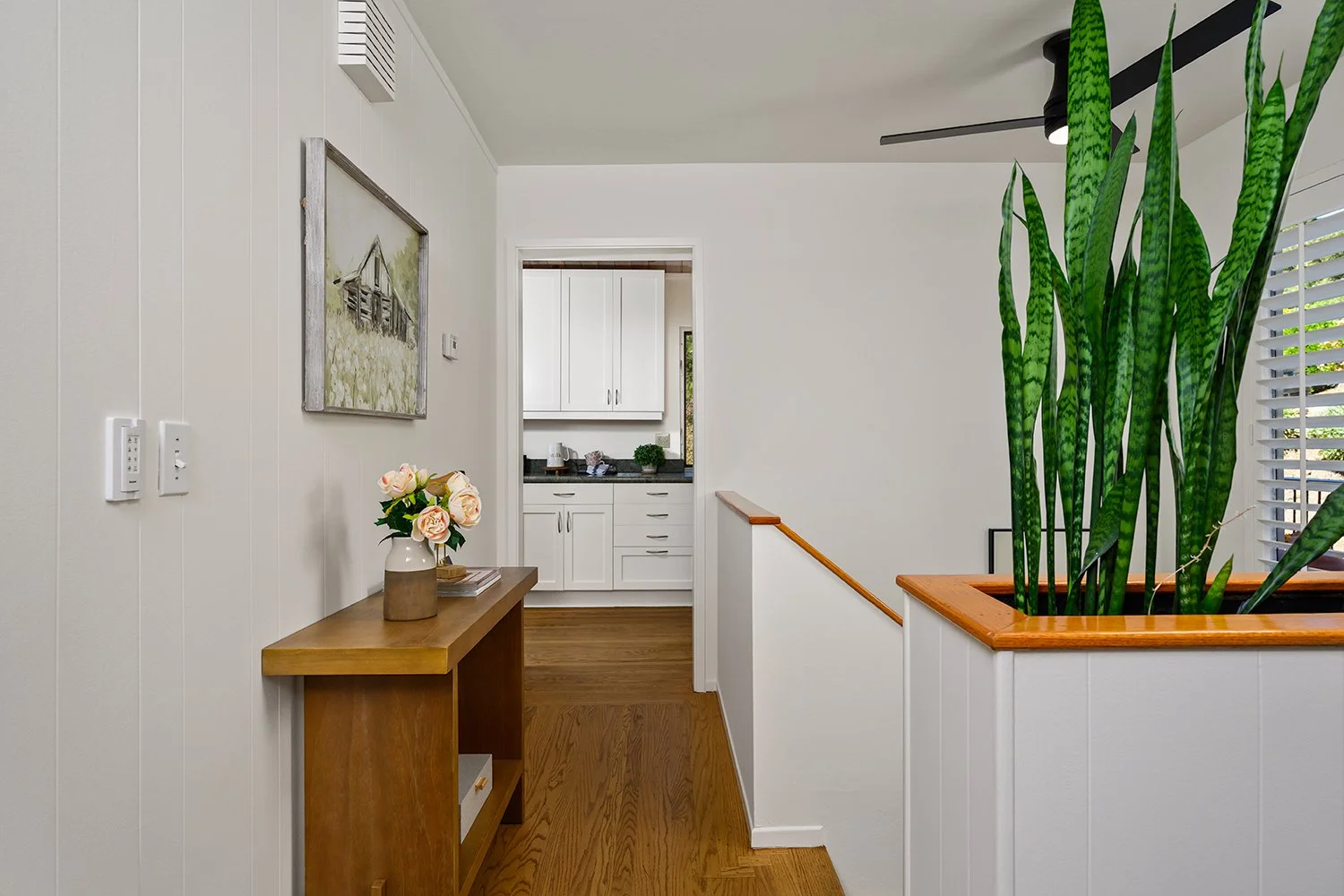 Interior view of a home with a wooden console table holding a vase of pink roses, located on a wall with white beadboard paneling, and a hall leading to a kitchen with white cabinets and black countertops. There is a large green snake plant in a wood