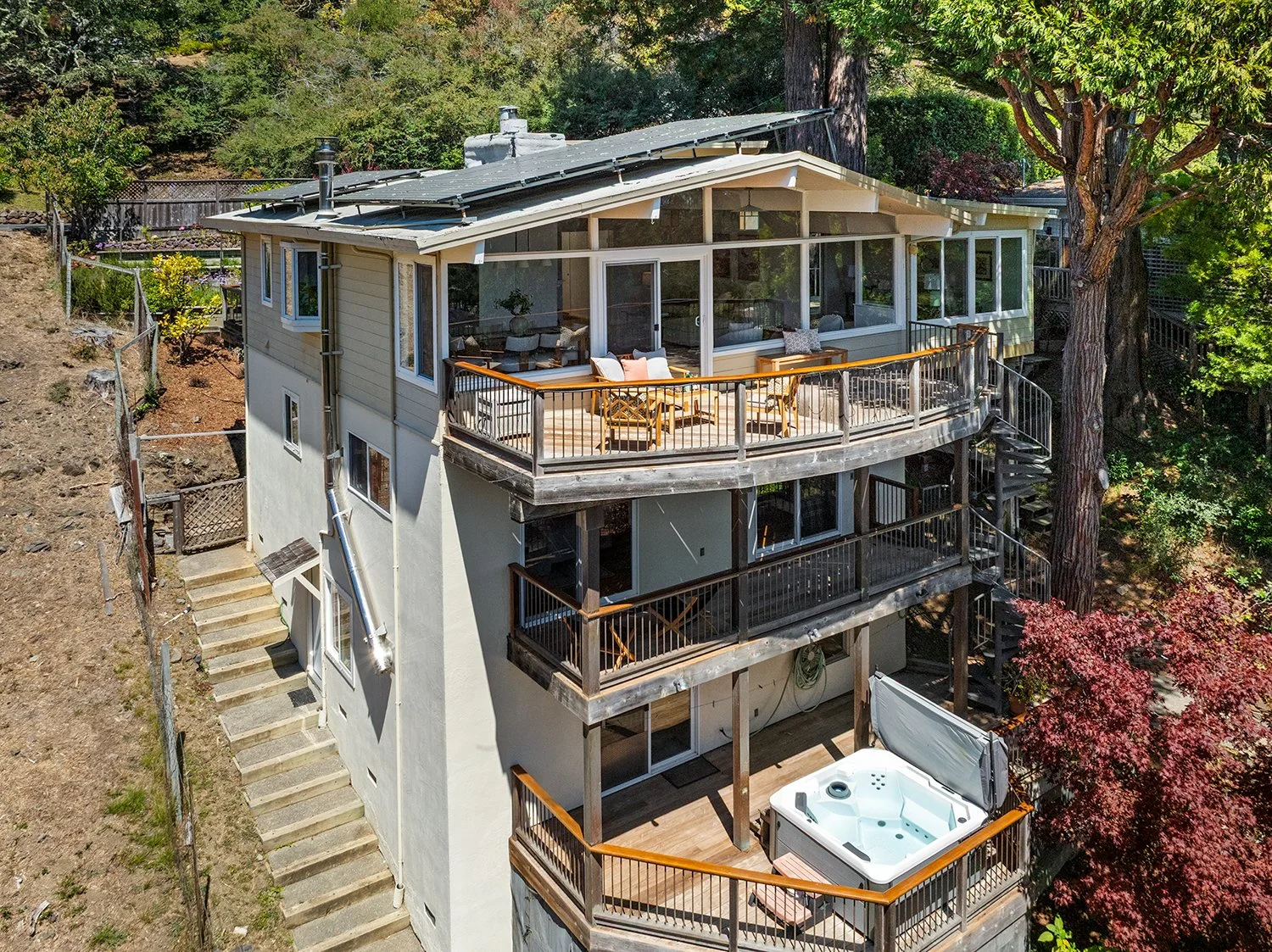 Multi-story house with multiple balconies, outdoor seating, hot tub, and stairs on hillside surrounded by trees and greenery.