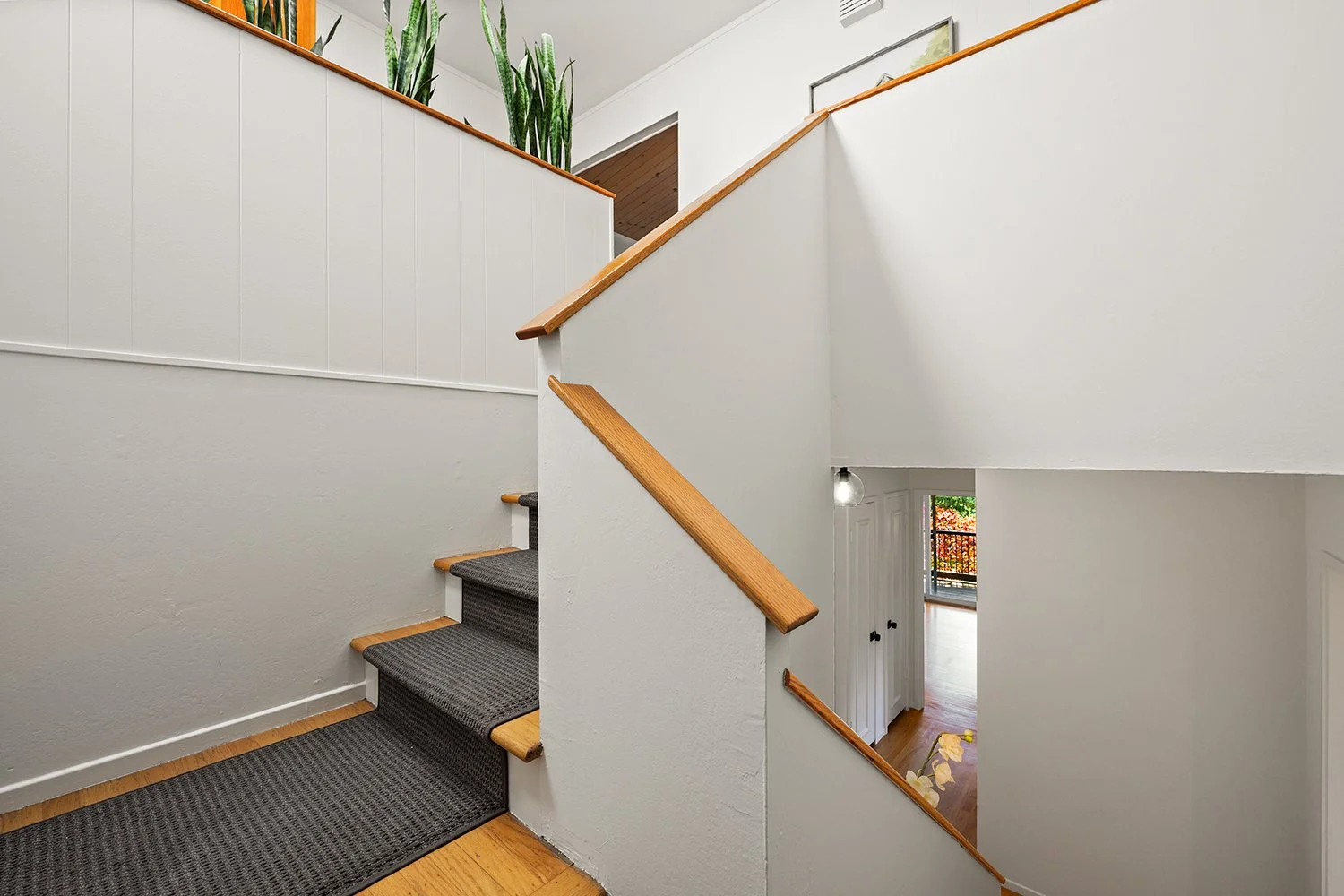 Interior of a residential staircase with wood handrails, gray carpeted steps, white walls, and a view of a doorway leading to a room with a balcony and outdoor scenery.