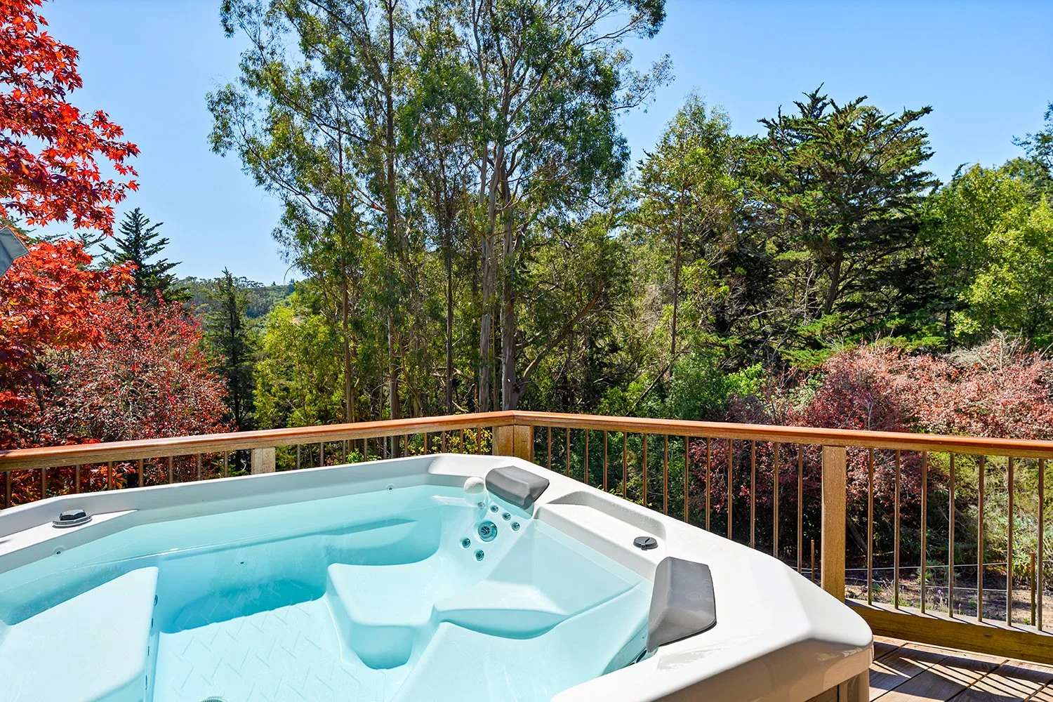 Hot tub on a wooden deck overlooking a lush, green forest with trees and flowering shrubs under a clear blue sky.