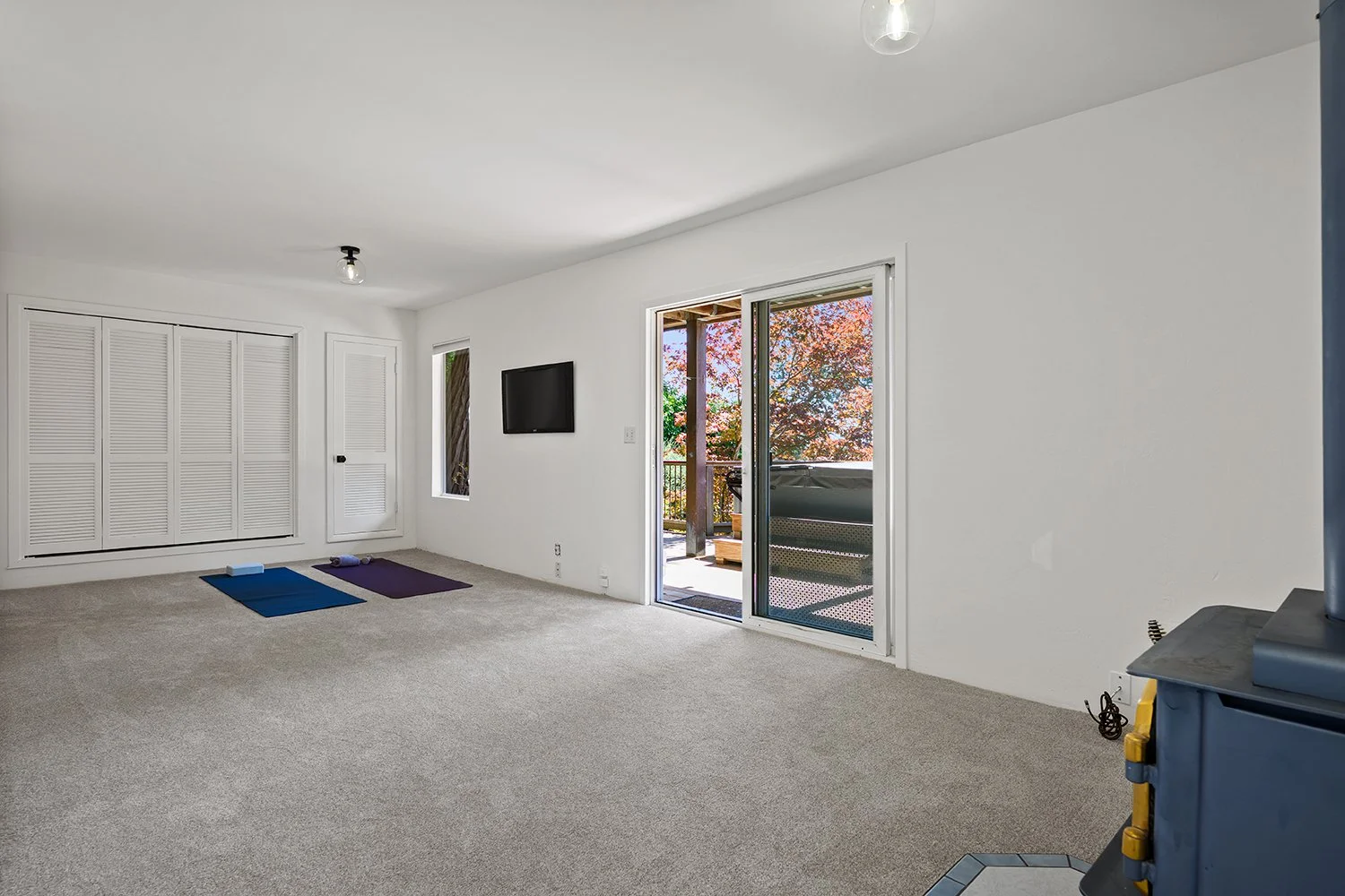 Empty living room with beige carpet, white walls, sliding glass door leading to a balcony, and a wall-mounted TV. Two yoga mats and props are on the floor, and a large window with fall foliage outside. Part of a black stove or heater in the foregroun