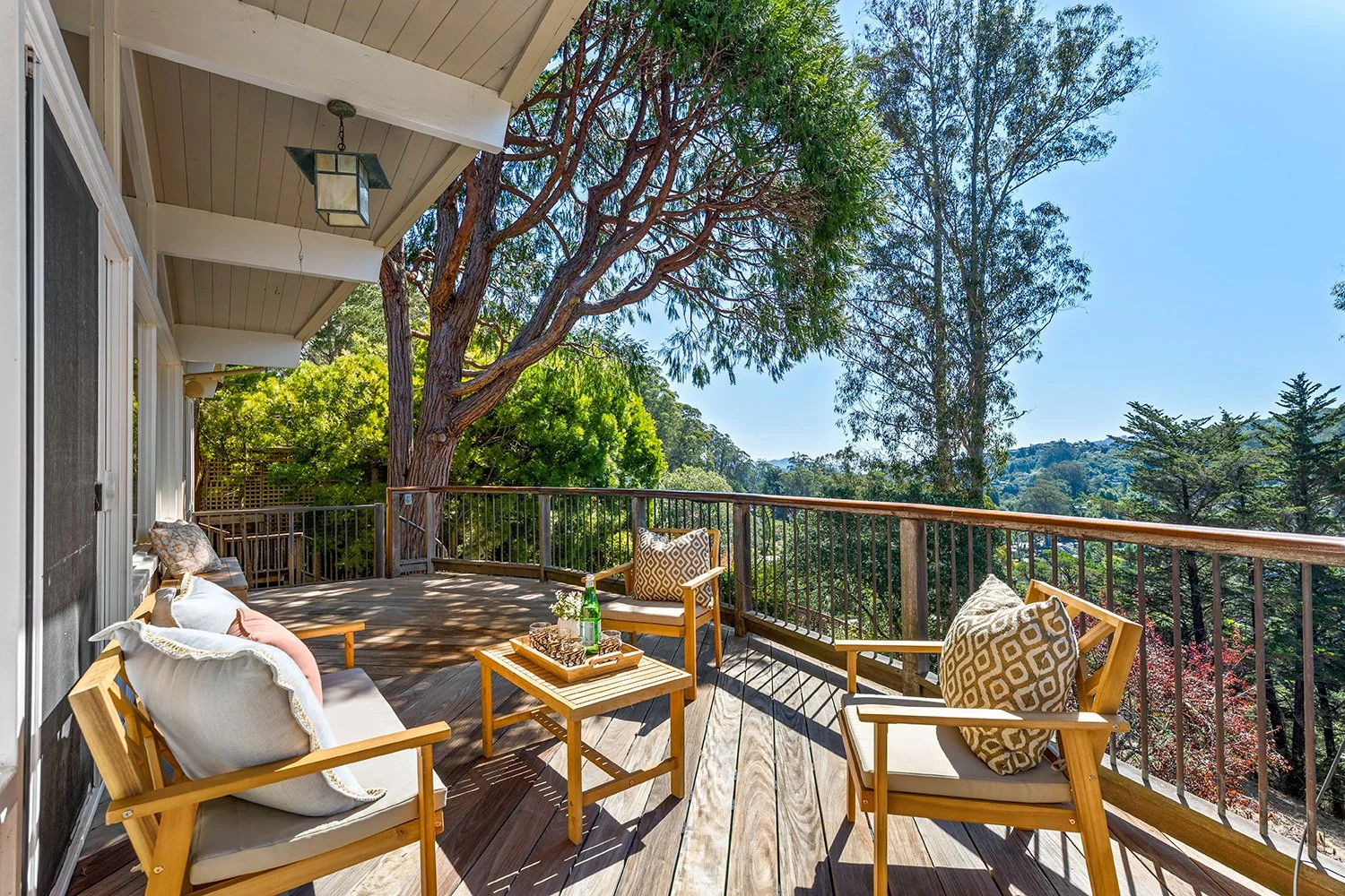 A wooden balcony with outdoor seating, including chairs with cushions, a small table with drinks, overlooking trees and a scenic landscape.