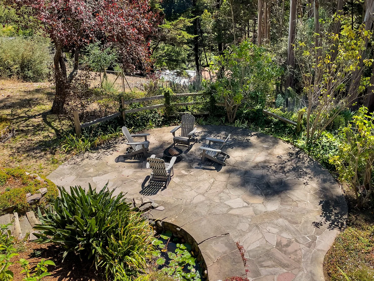 A circular stone patio with five Adirondack chairs arranged around a fire pit, surrounded by lush green plants and mature trees in a garden setting.