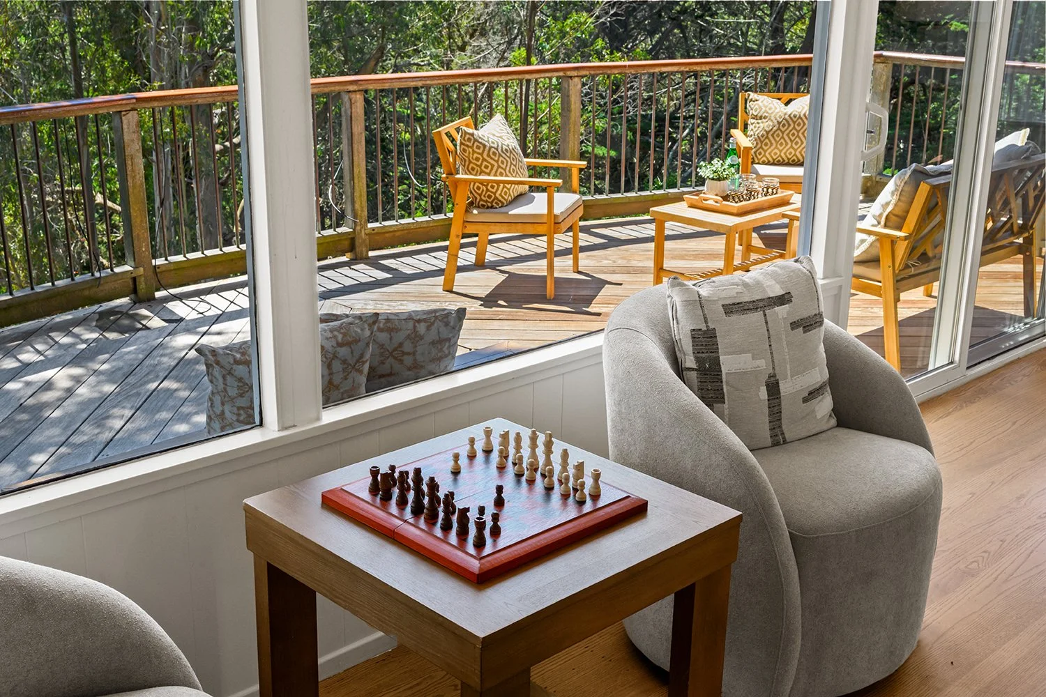 Living room with a chessboard on a small wooden table, beige armchair with patterned pillow, large glass sliding doors leading to a wooden balcony with outdoor chairs and cushions, trees and greenery outside.