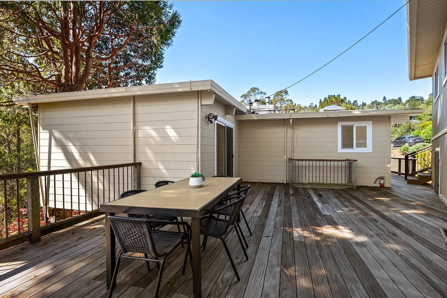 Wooden deck with outdoor dining table and six chairs, beige house with sliding glass door, small window, greenery, trees, parked car in the background, and clear blue sky.