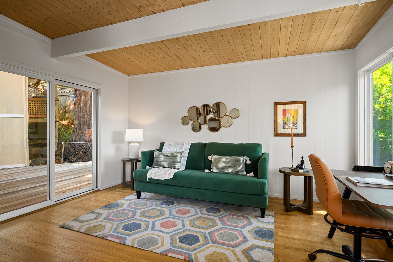 Living room with green sofa, colorful rug, wooden floors, sliding glass door to outdoor deck, white walls, wood ceiling, side tables, lamp, wall art, and a desk with an office chair.