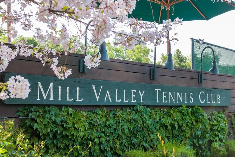 Sign reading 'Mill Valley Tennis Club' mounted on a wooden fence, surrounded by green ivy and pink flowering tree branches.