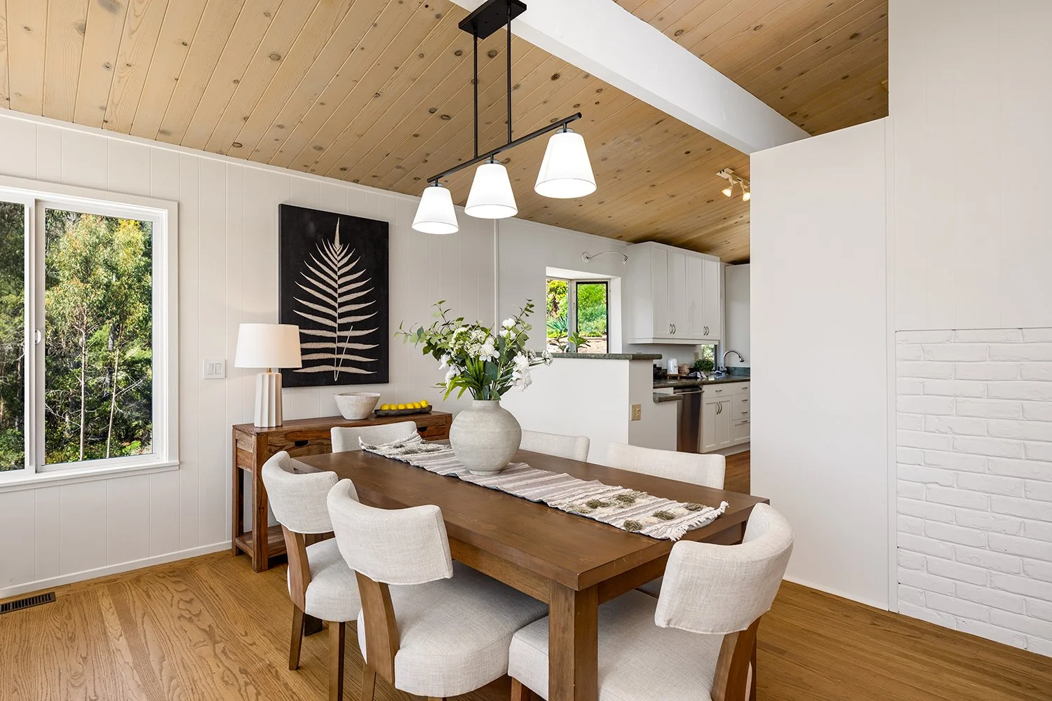 A dining room with a wooden table surrounded by six beige upholstered chairs, a large vase with flowers on the table, and a window showing greenery outside. The room has white walls, wooden ceiling, and hardwood floors. There's a black and white leaf