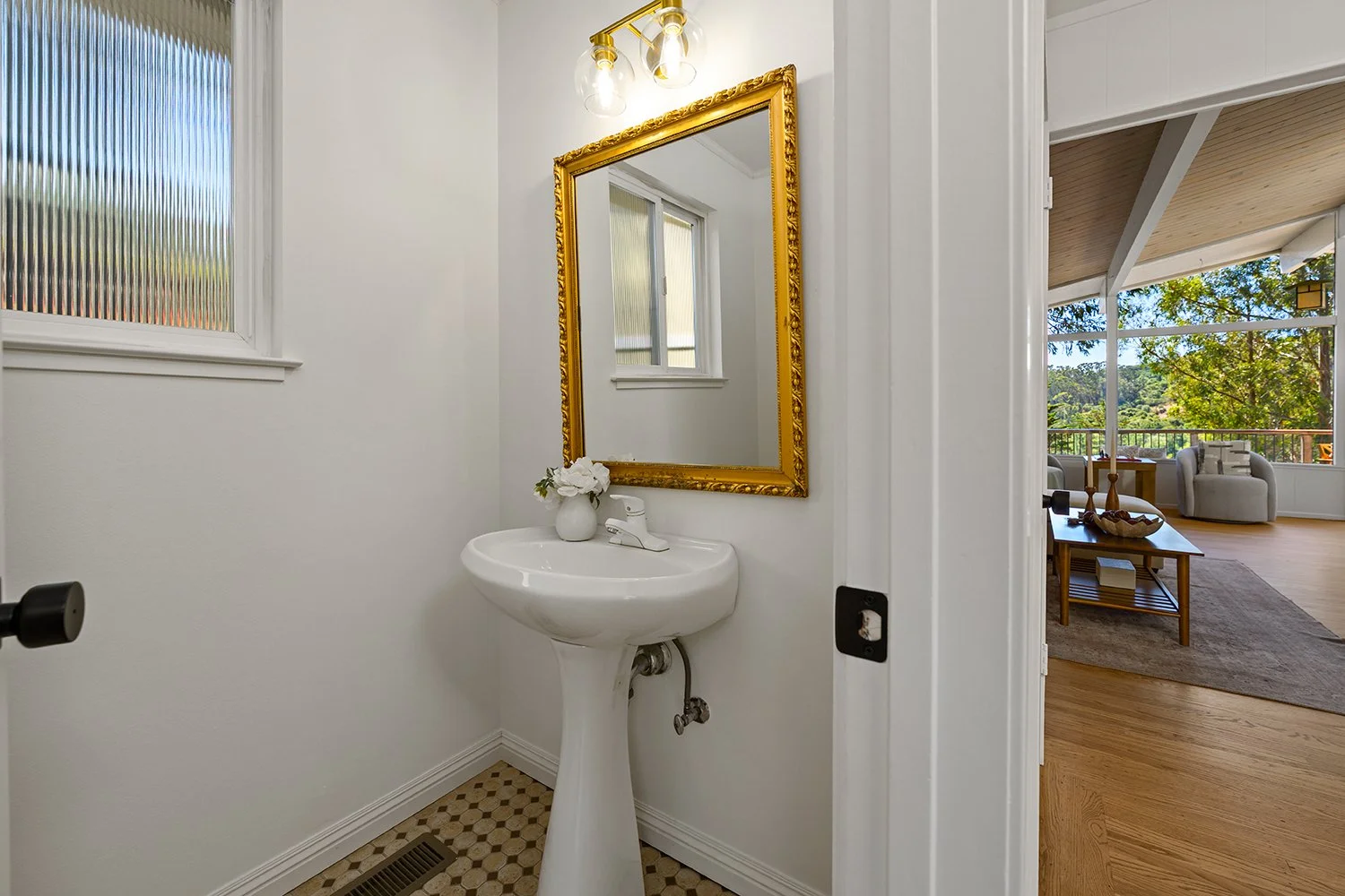 A small white bathroom with a pedestal sink, a vase with white flowers, a gold-framed mirror, and a frosted window. The bathroom is adjacent to a bright living room with large windows and wooden floors.