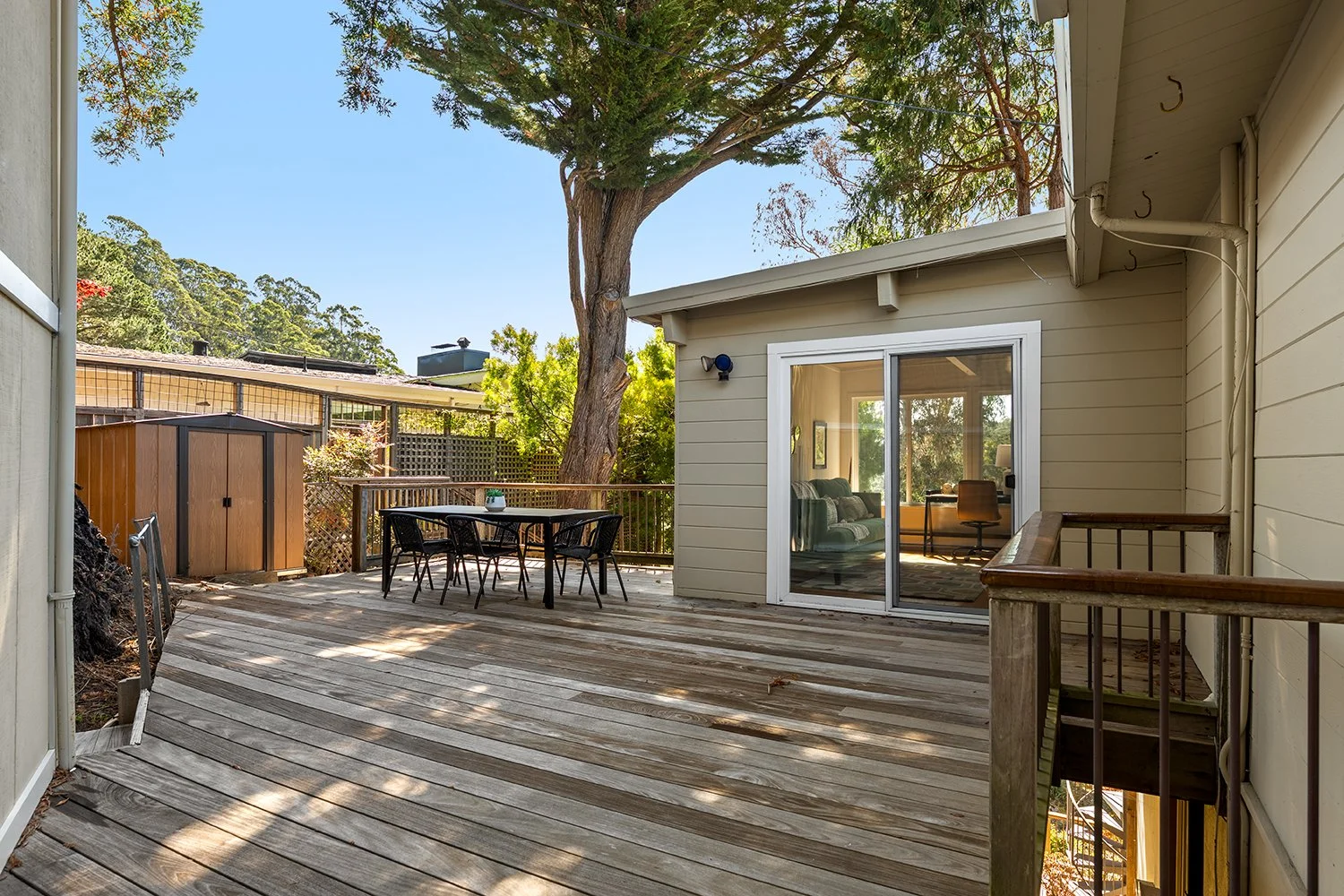View of a wooden outdoor deck adjacent to a yellow house with a sliding glass door. The deck has a black outdoor dining table with five chairs, a small shed, and surrounding trees providing shade.