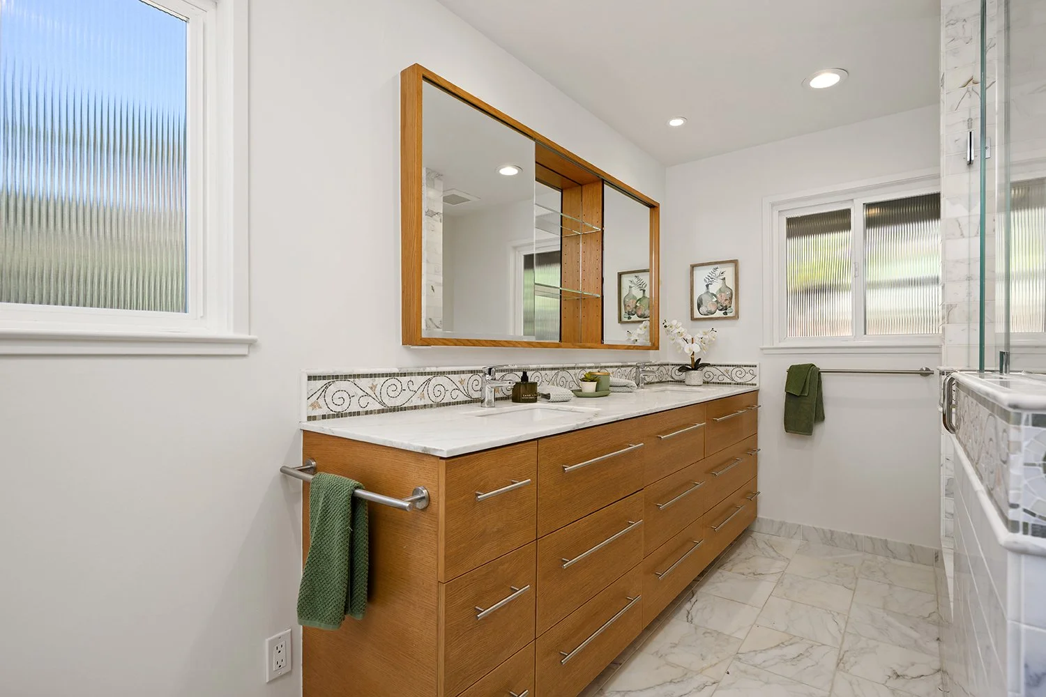 Modern bathroom with wooden vanity, marble countertop, large mirror, two windows with frosted glass, and decorative tile border