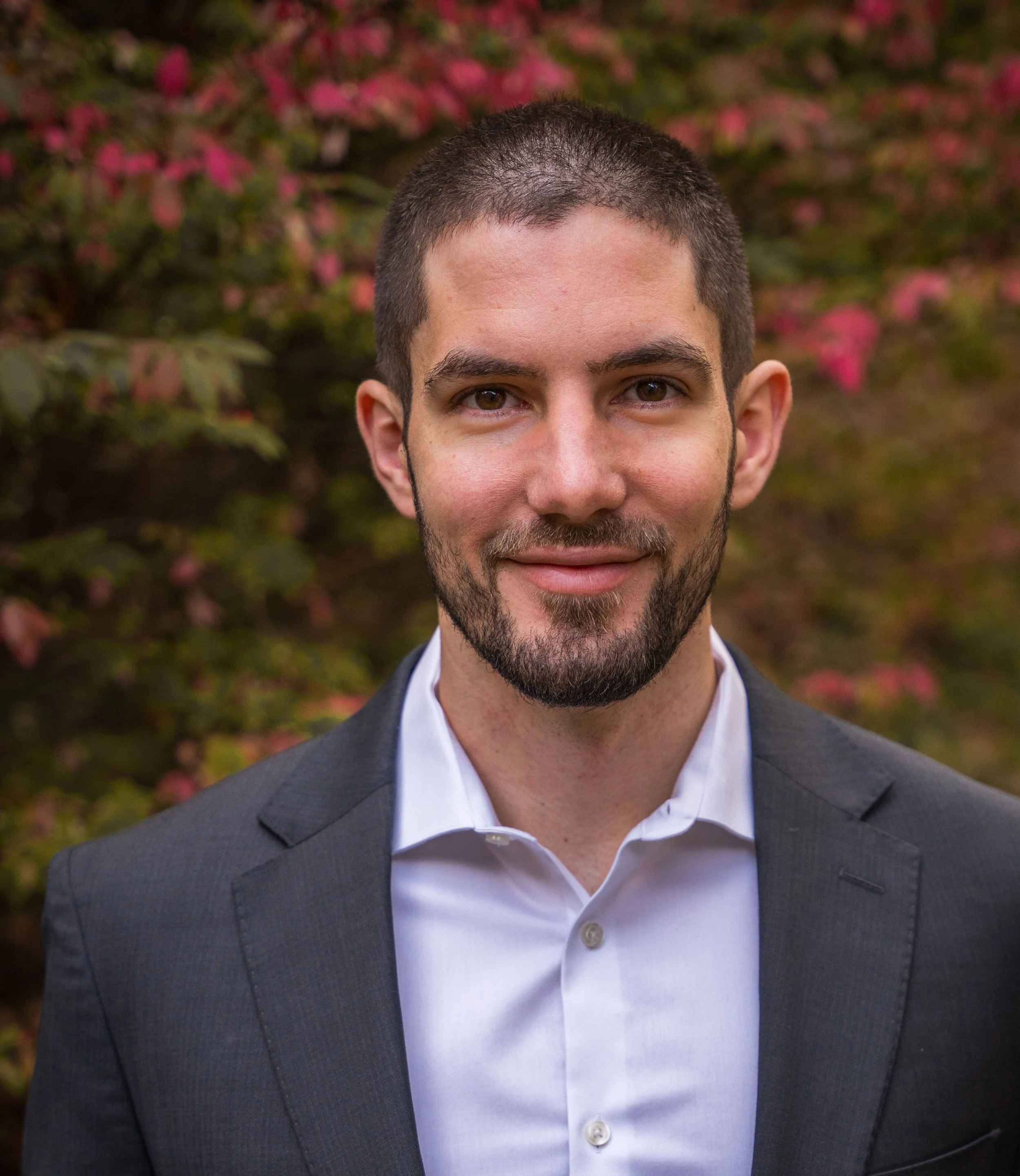 Close-up of a Dr. Joshua Neer, founder of Redeeming Motion, wearing a dark gray suit and white shirt, standing outdoors with pink flowers and foliage in the background.