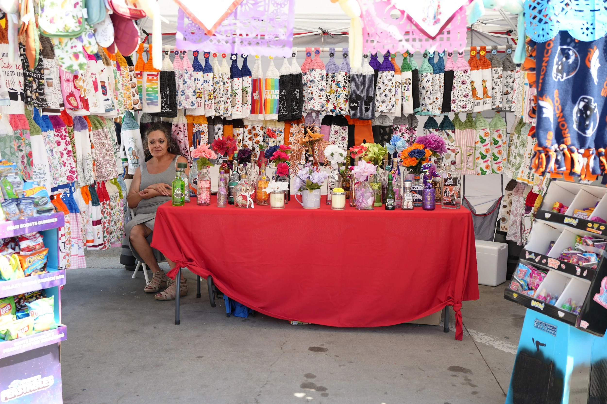 A woman sitting behind a red tablecloth-covered table at a booth selling colorful handmade sock puppets, decorated jars with flowers, and bottles of drinks at an outdoor market. The background displays hanging socks and other merchandise.