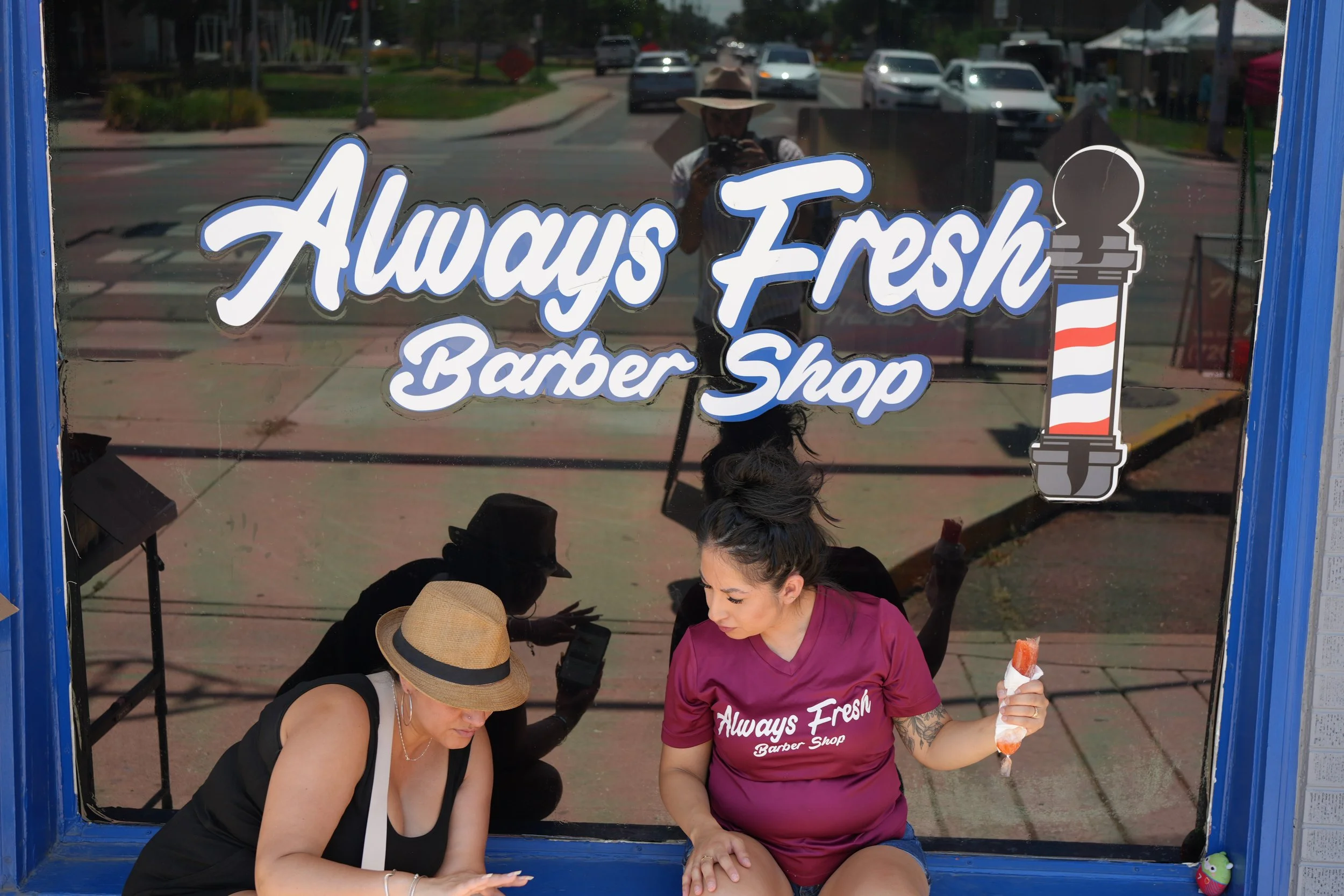 Two women, one in a black top and another in a pink barber shirt, sitting inside a blue barber shop window, with one of them holding an ice cream cone. The reflection of a person with a hat is visible on the window behind them.