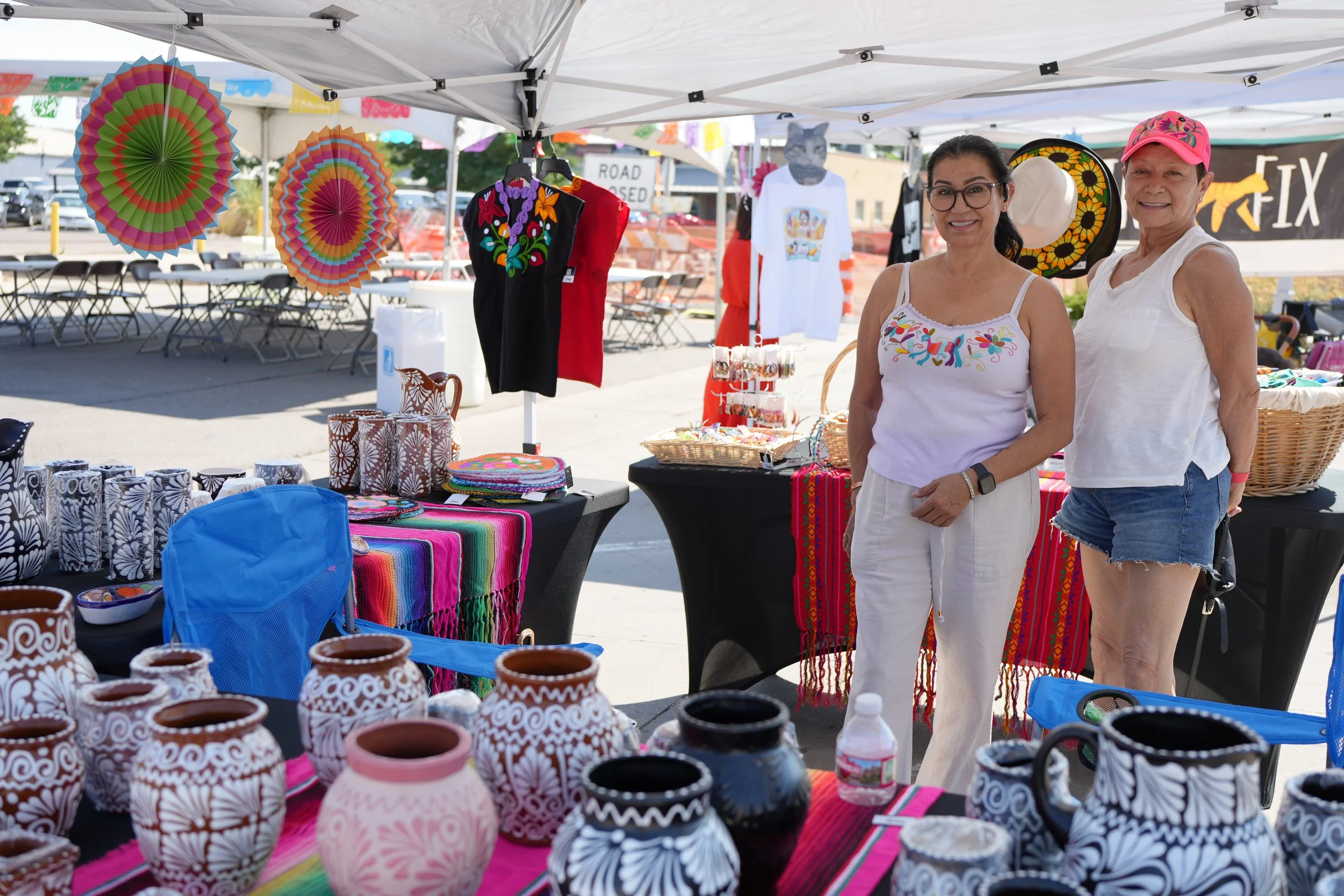 Two women standing behind a table at a marketplace booth, displaying colorful pottery, textiles, and accessories. The booth has hanging paper decorations and embroidered clothing.