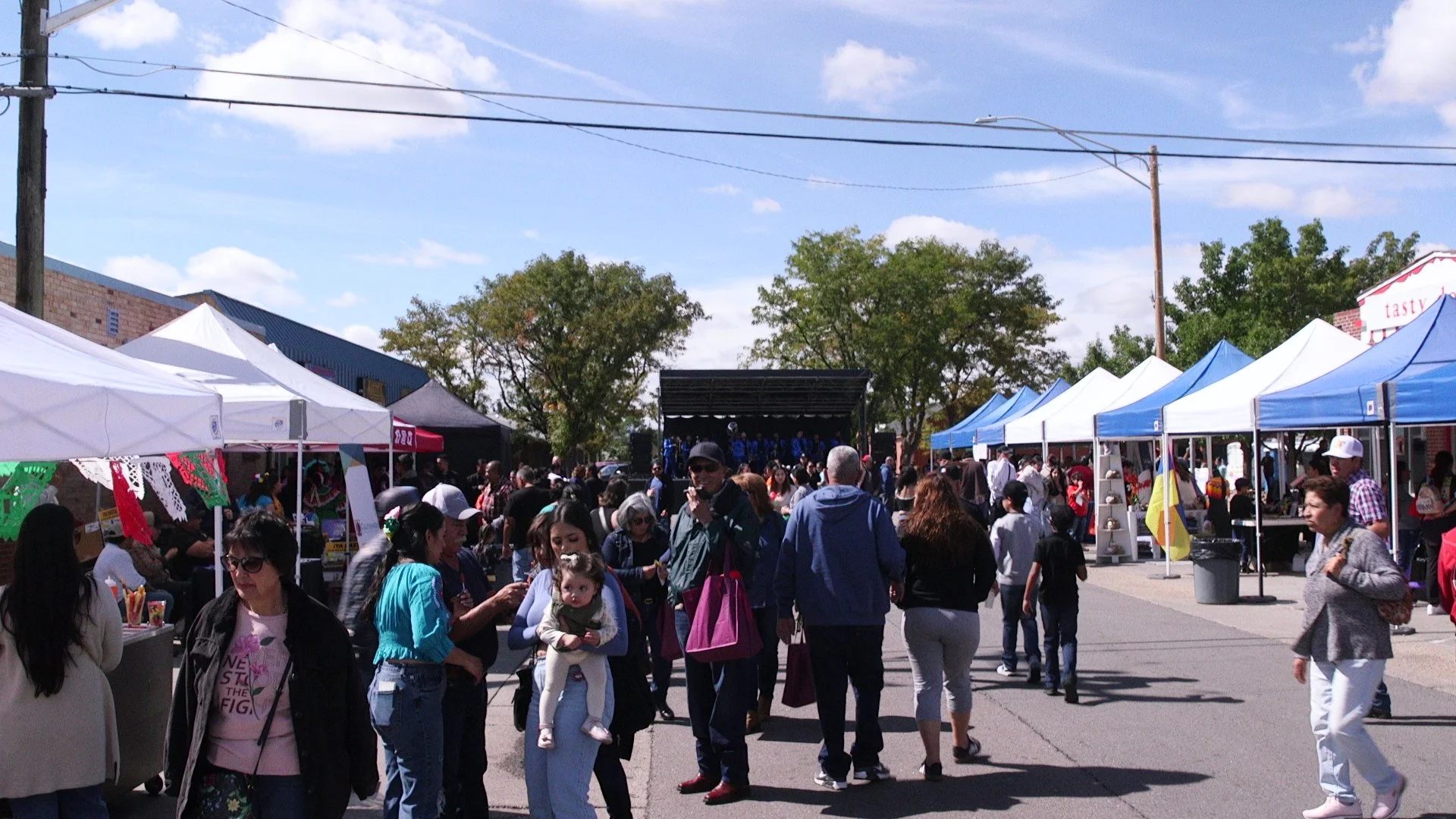 People attending outdoor market with vendor tents and a stage in the background under a partly cloudy sky.