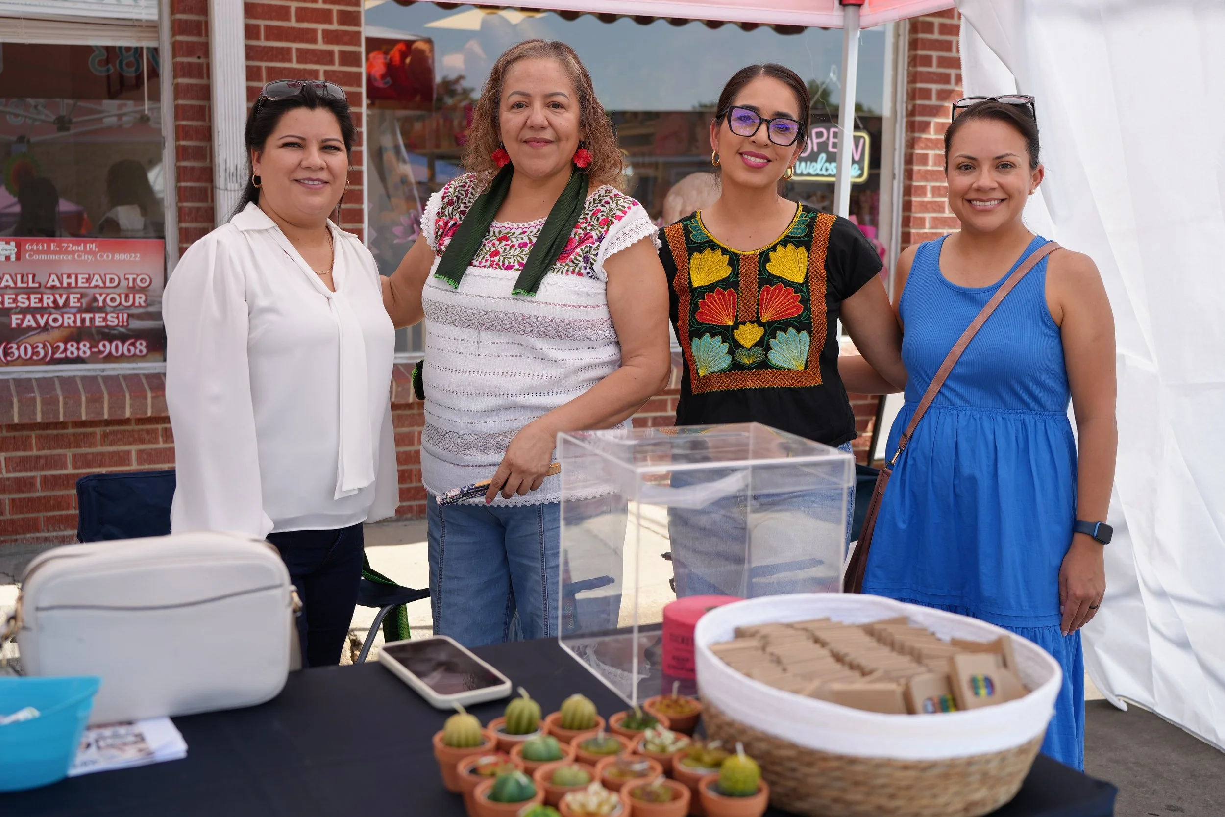 Four women standing behind a table at an outdoor market or fair, smiling at the camera. The table displays small potted plants, cookies, and other baked goods. The background includes a brick building and a tent.