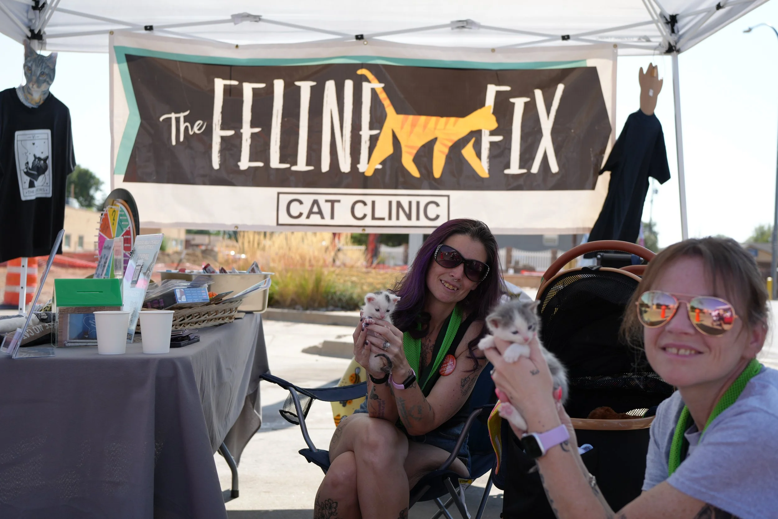 People seated at a booth labeled the Feline FX Cat Clinic, holding small kittens. The booth has a table with informational materials and two mannequins with cat-themed costumes and clothing. There is a banner with a stylized cat silhouette hanging ab