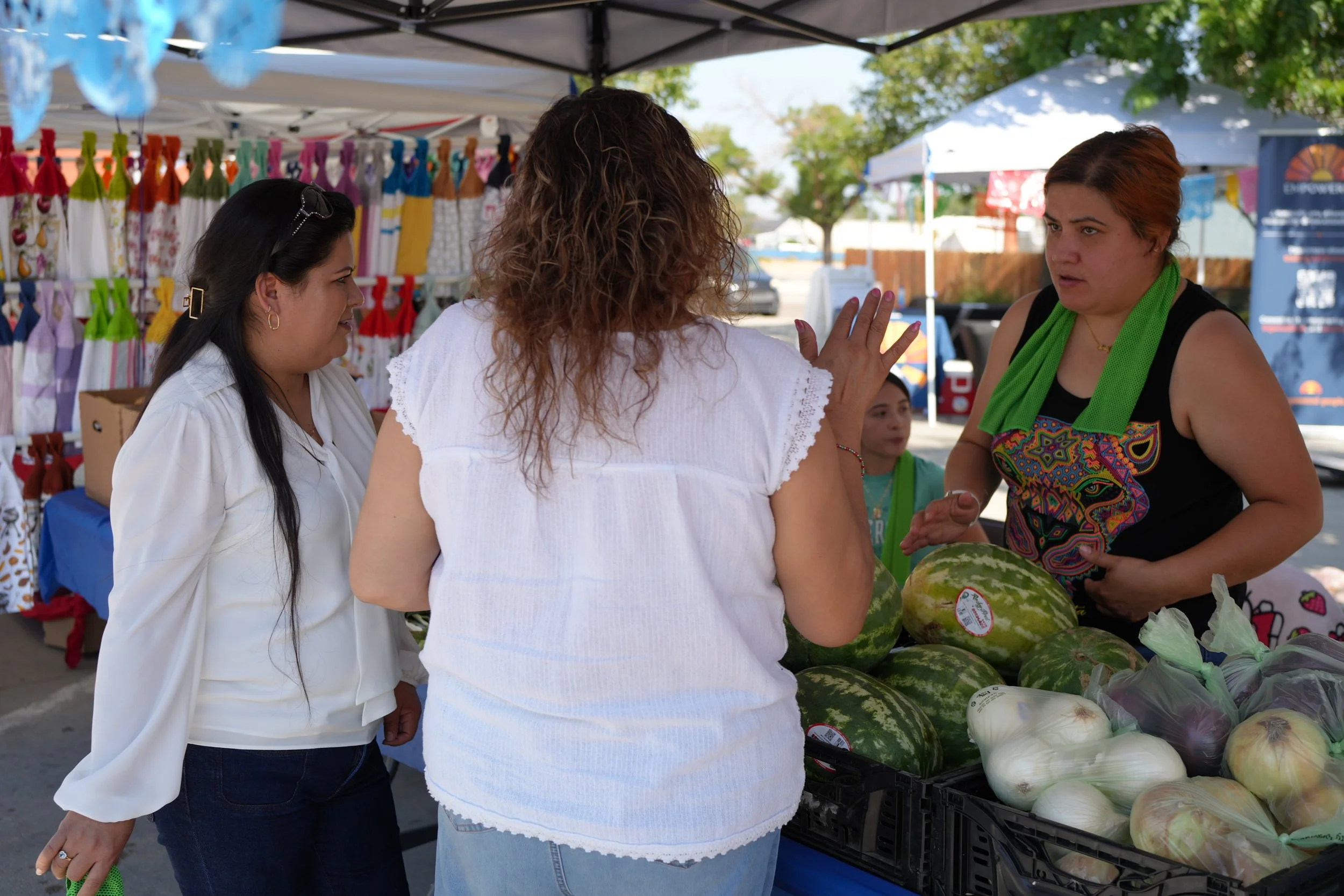 Three women are talking at a farmers market stand selling watermelons and onions, with colorful cloth bags hanging in the background and tents set up under trees.