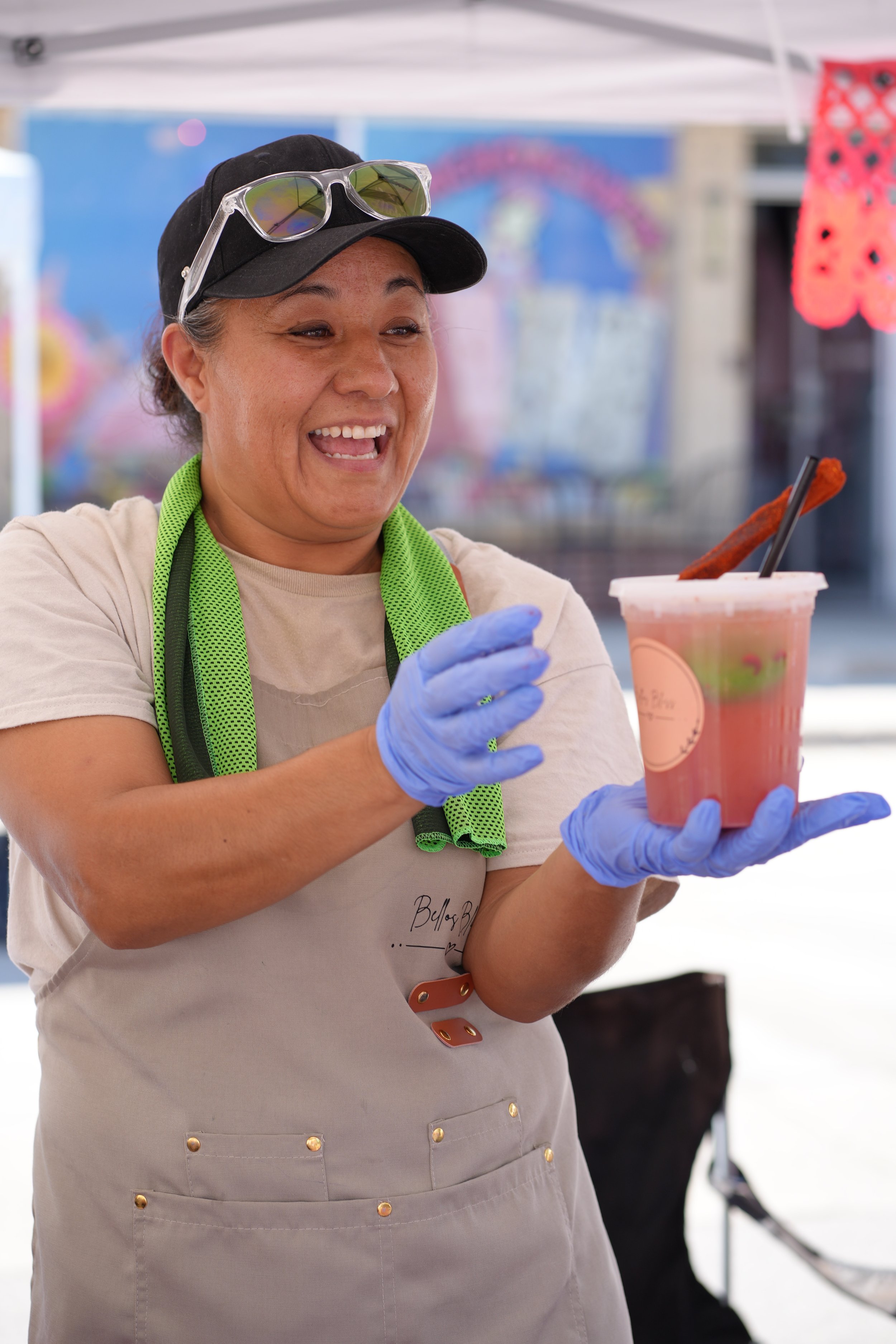A woman in a beige dress with gold buttons, a black baseball cap, and sunglasses on her head, holding a pink drink with a black straw and a stick in it at an outdoor event.