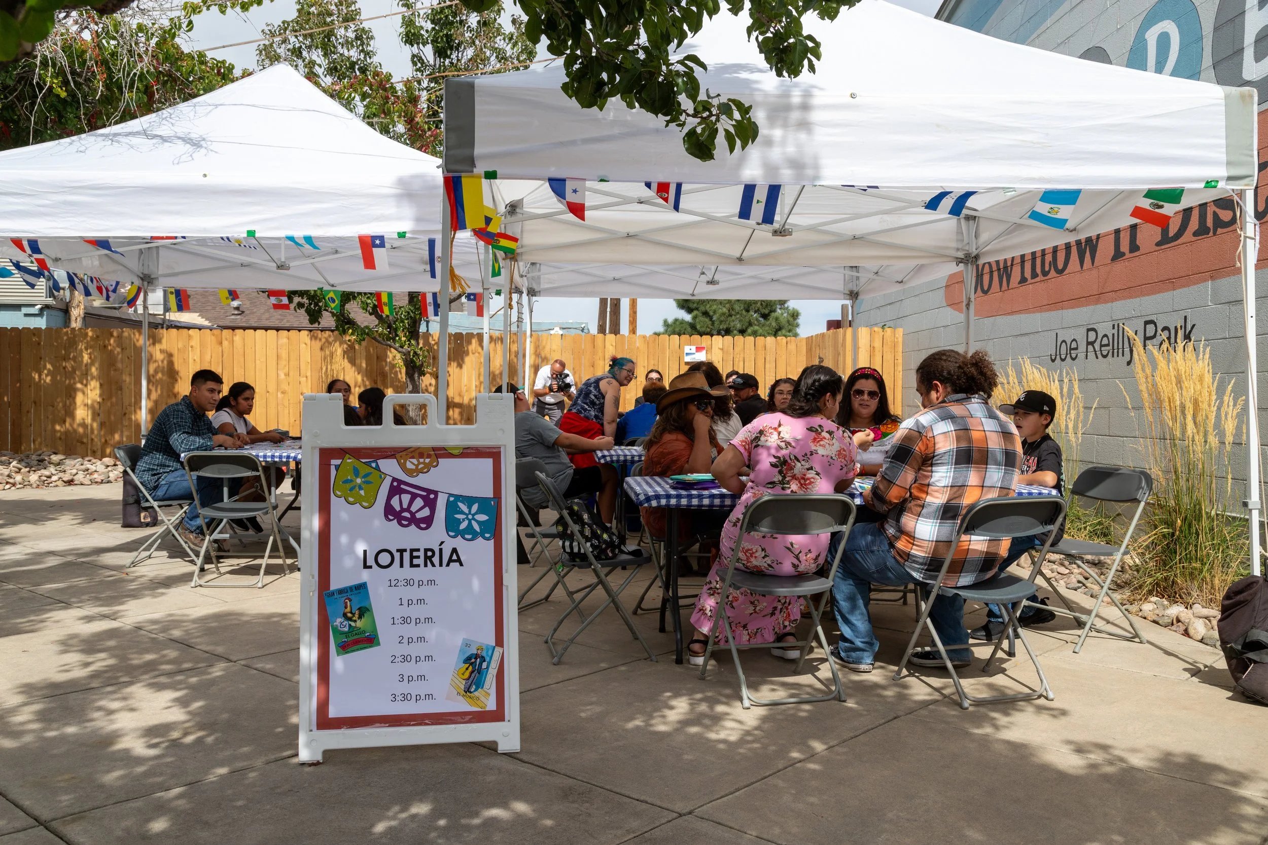 People sitting at tables under white tents decorated with colorful international flags at an outdoor event. A sign in the foreground displays a schedule for a lottery on a sunny day.