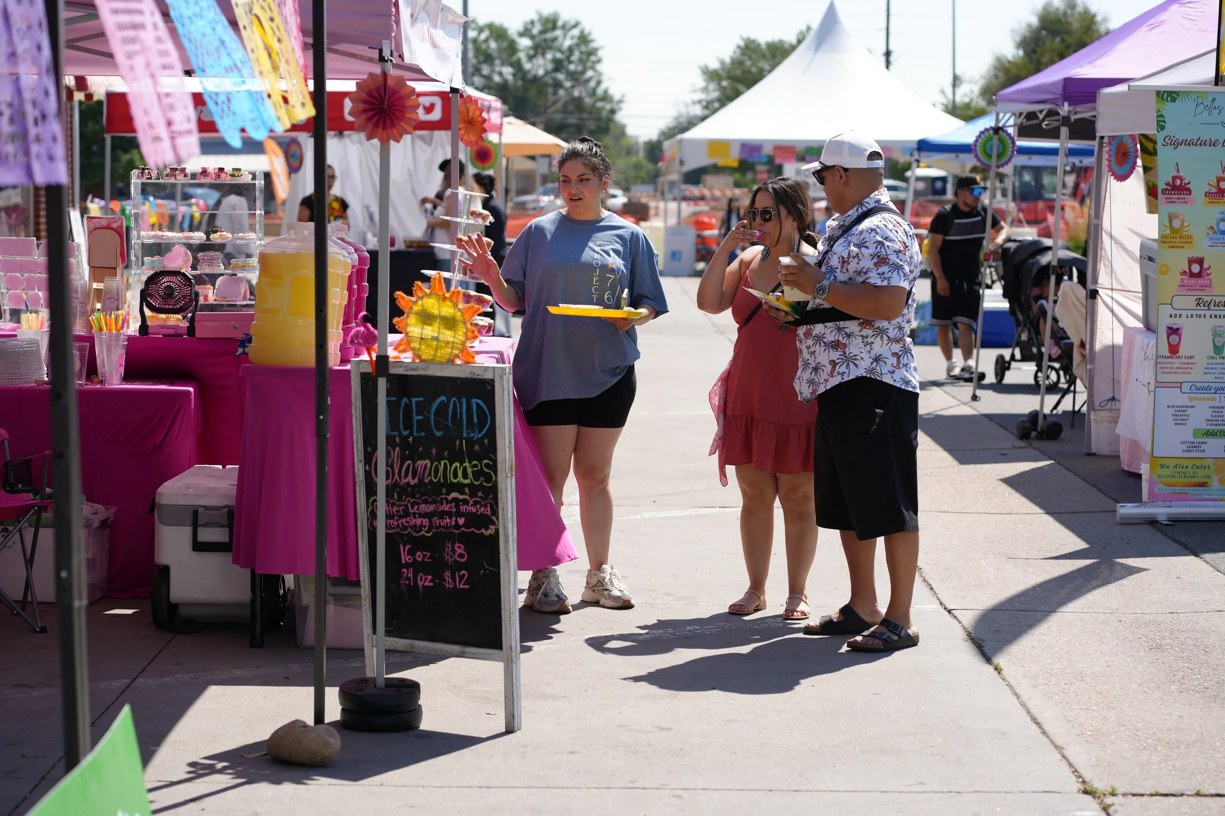 A scene at an outdoor market with tents and stalls. Three people are looking at a brightly decorated stall selling souvenirs and trinkets. The woman in the center is wearing a gray T-shirt, holding a yellow tray, and gesturing with her hand. The woma