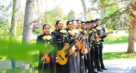 A group of seven musicians dressed in traditional black attire with gold accents, holding guitars and violins, standing outdoors in a park with green trees and grass.