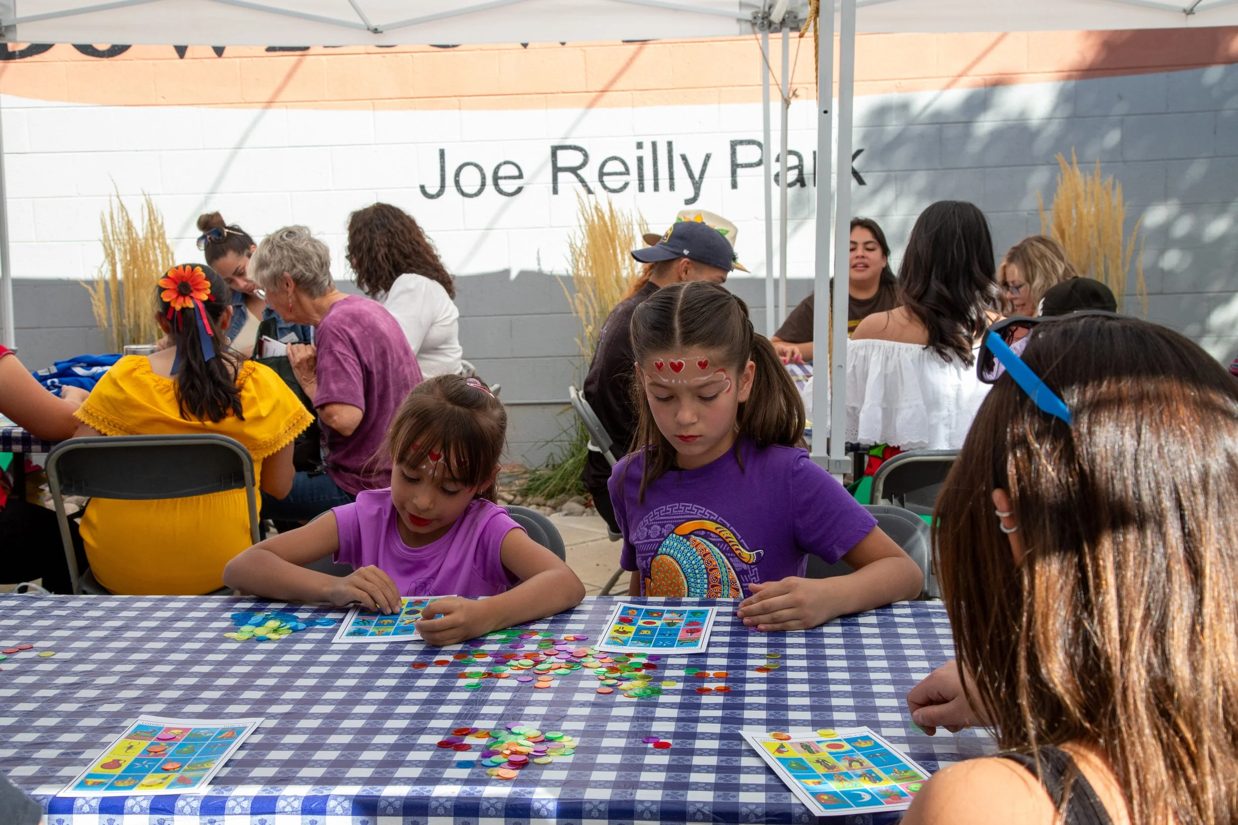 People sitting at a table playing a colorful board game with cards and tokens, outdoors under a canopy with a wall in the background and the text 'Joe Reilly Park' on it.