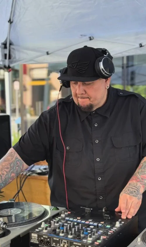 A male DJ in black clothing and headphones playing music at an outdoor event under a tent.