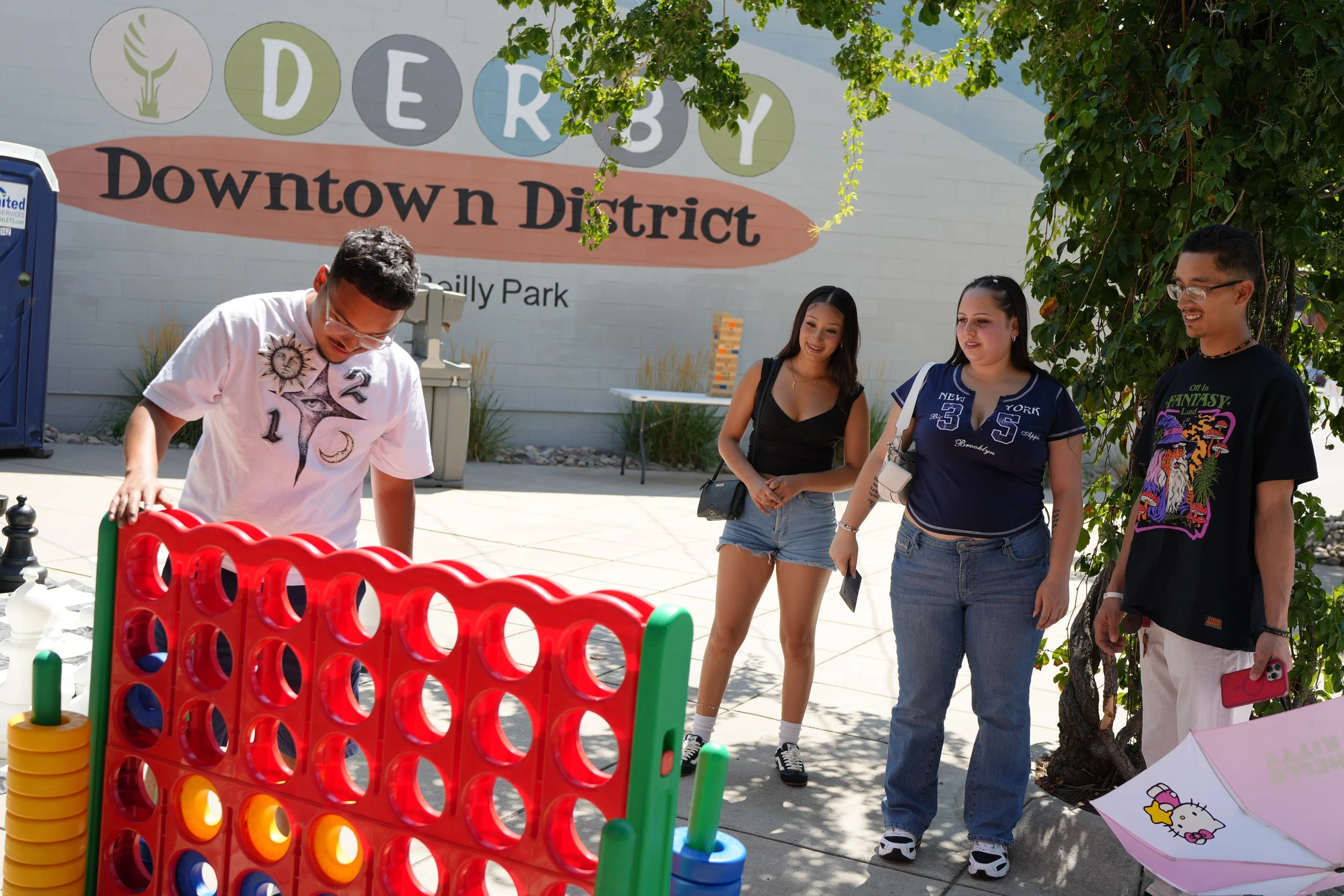 A group of four young adults playing Connect Four outdoors near a sign that reads 'DERBY Downtown District.' The young man in a white shirt is about to make a move in the game, while the three others, two women and one man, watch with smiles. There a