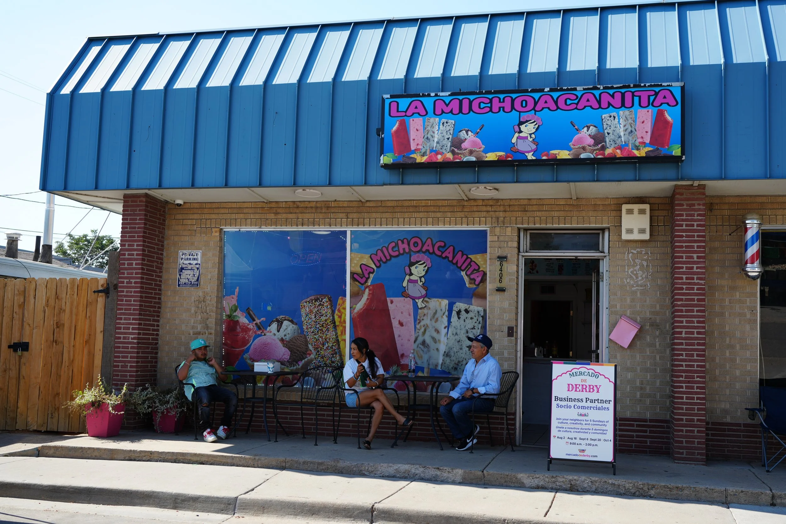 Ice cream shop with bright colorful signage, people sitting outside, and a sidewalk sign advertising community events.