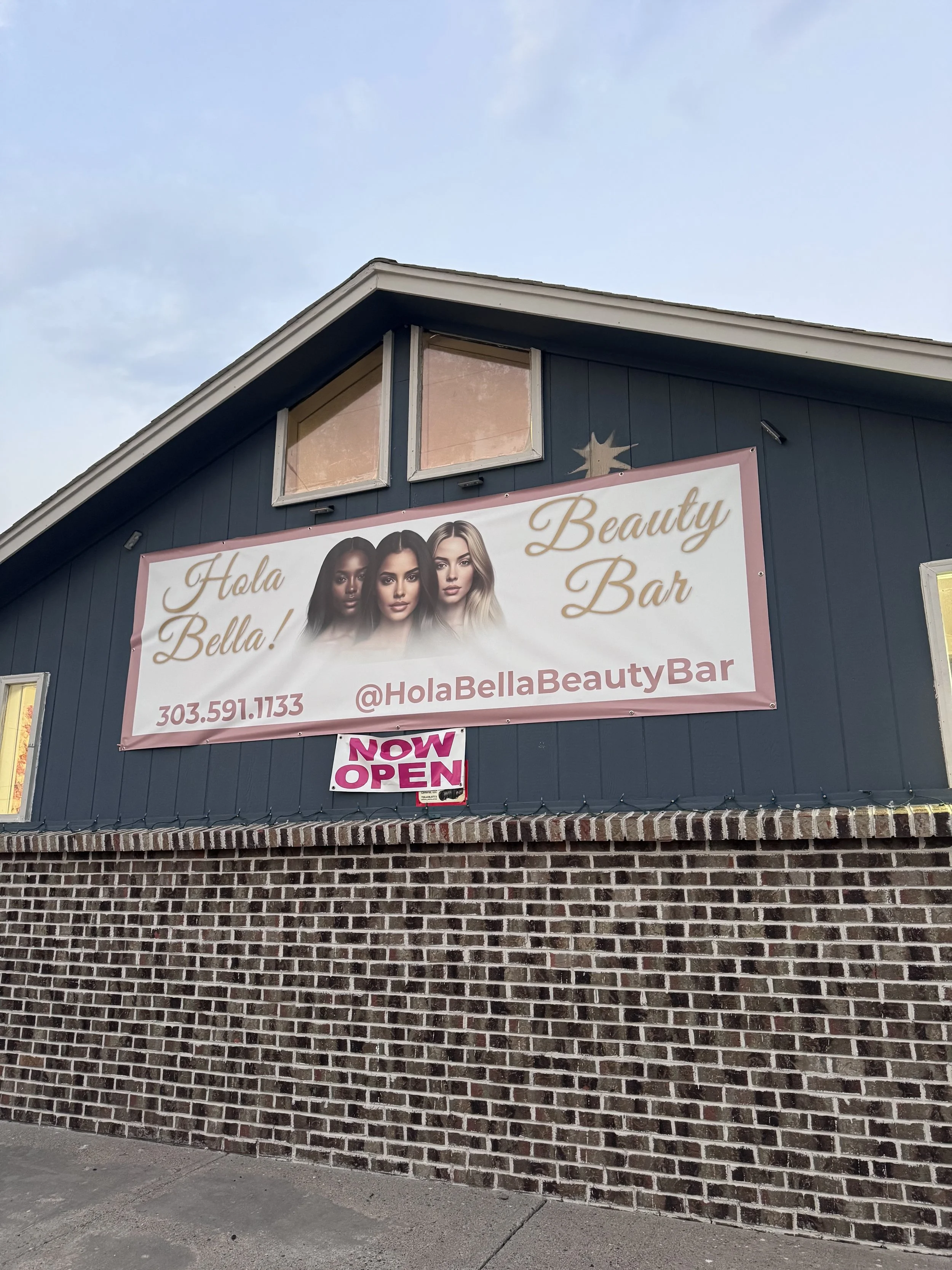 A black building with a brick foundation and two windows near the roof. A large banner advertises Hola Bella Beauty Bar, featuring images of three women with different hair types. A small pink sign reads 'NOW OPEN' underneath.