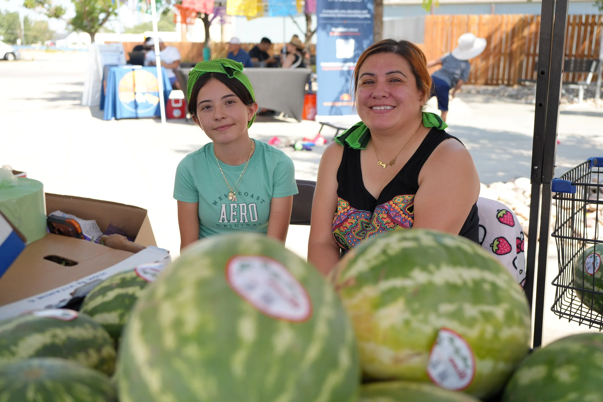 A woman and a girl smiling behind a display of watermelons at an outdoor market or fair.