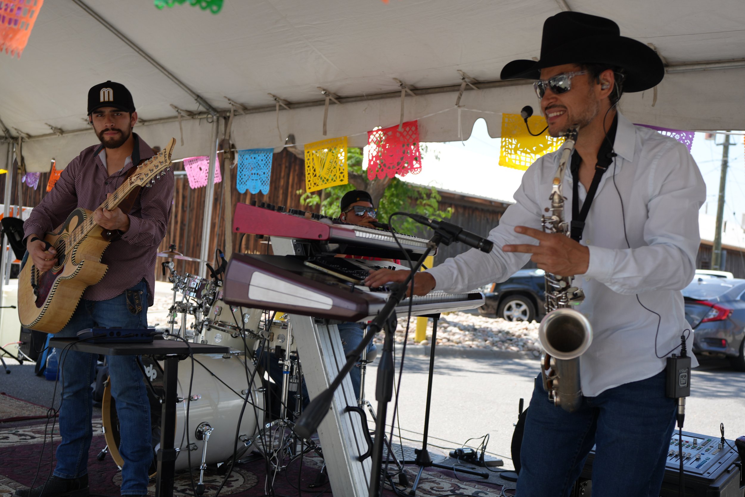 Musicians performing at an outdoor event, one playing guitar and another playing saxophone, with colorful papel picado banners hanging above.