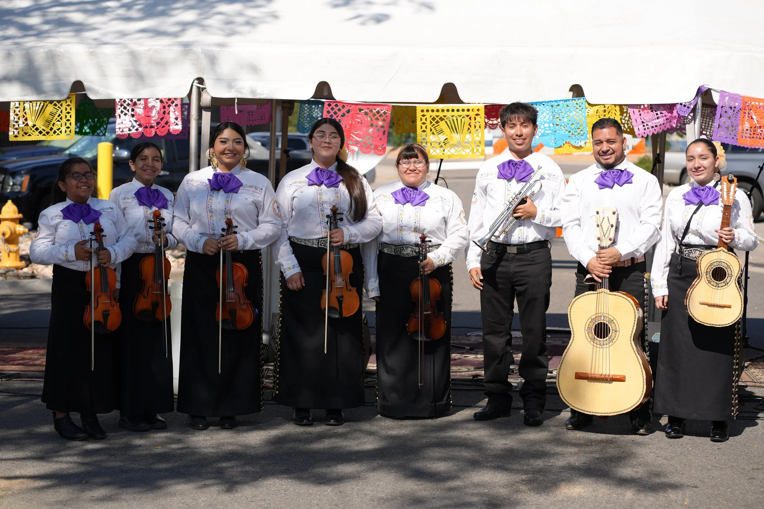 A group of nine musicians dressed in traditional Mexican clothing, standing in a line under a canopy decorated with colorful papel picado banners. Some of them are holding musical instruments including violins, guitars, and a trumpet.