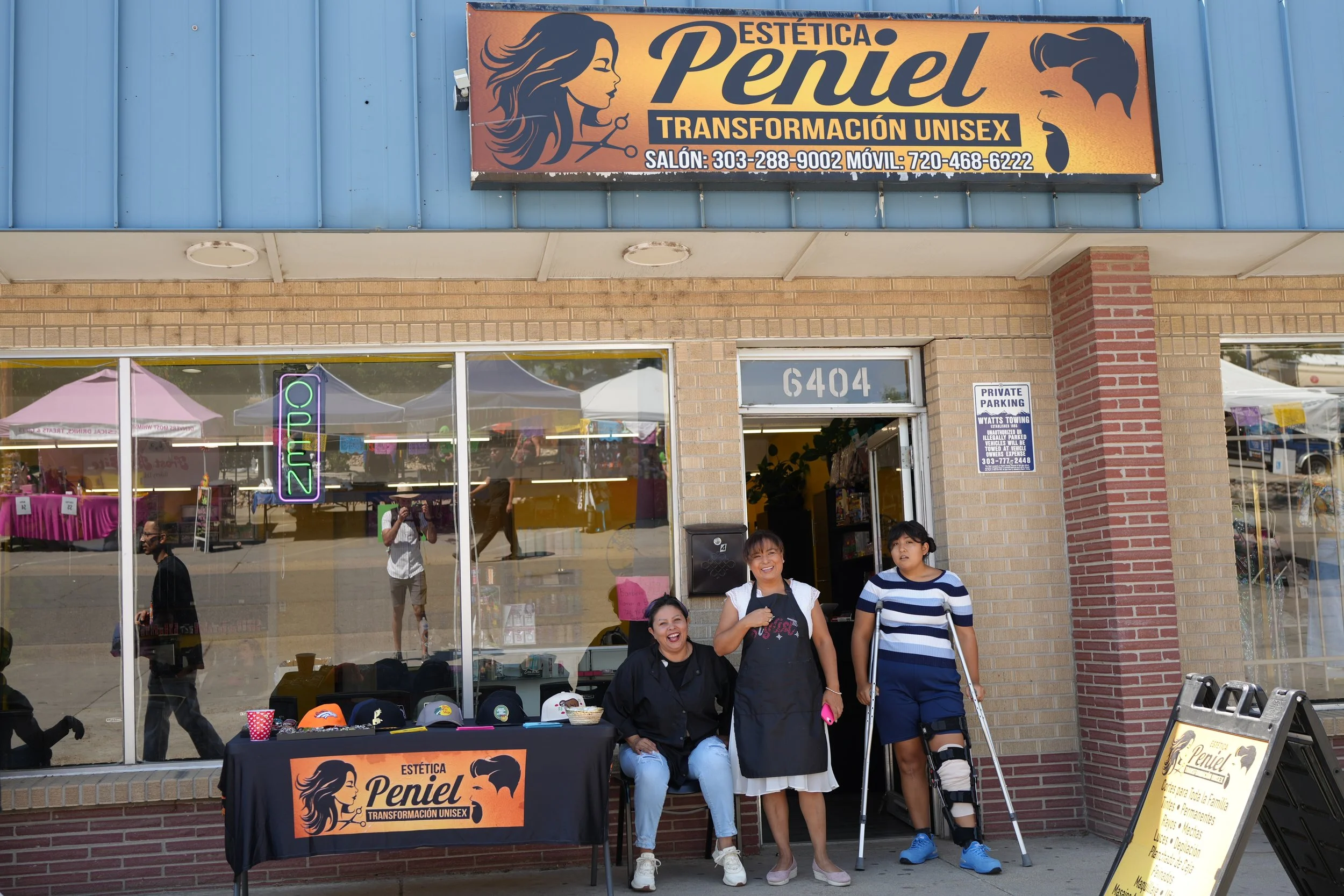 People standing outside a beauty salon named Estética Penel, with a sign advertising unisex transformation services, and a table with hats and a sign in front.