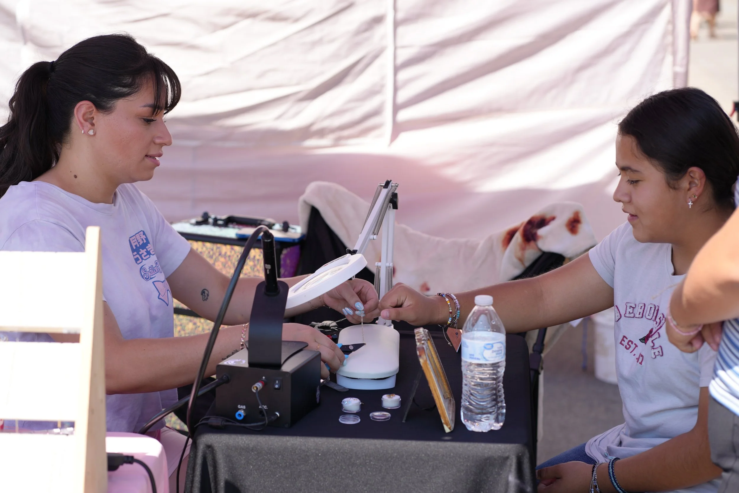 Two women sitting at a table outside, engaged in a nail or beauty treatment. One woman is receiving the treatment while the other is performing it. There are equipment and supplies on the table, including a water bottle and small containers. A person